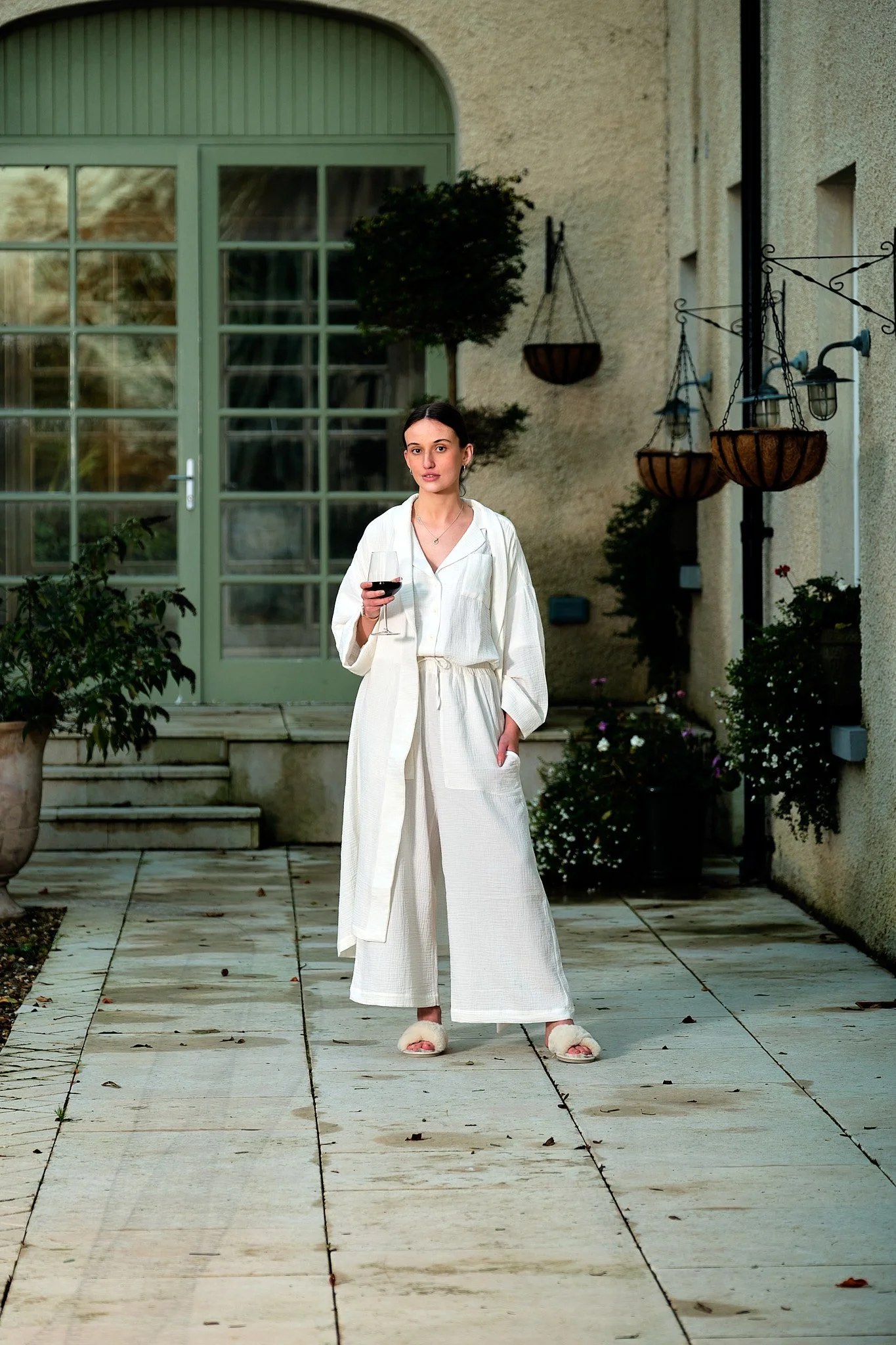A woman standing outdoors on stone patio holding a glass of red wine, dressed in a white robe, with potted plants, hanging baskets, and a large glass door behind her.