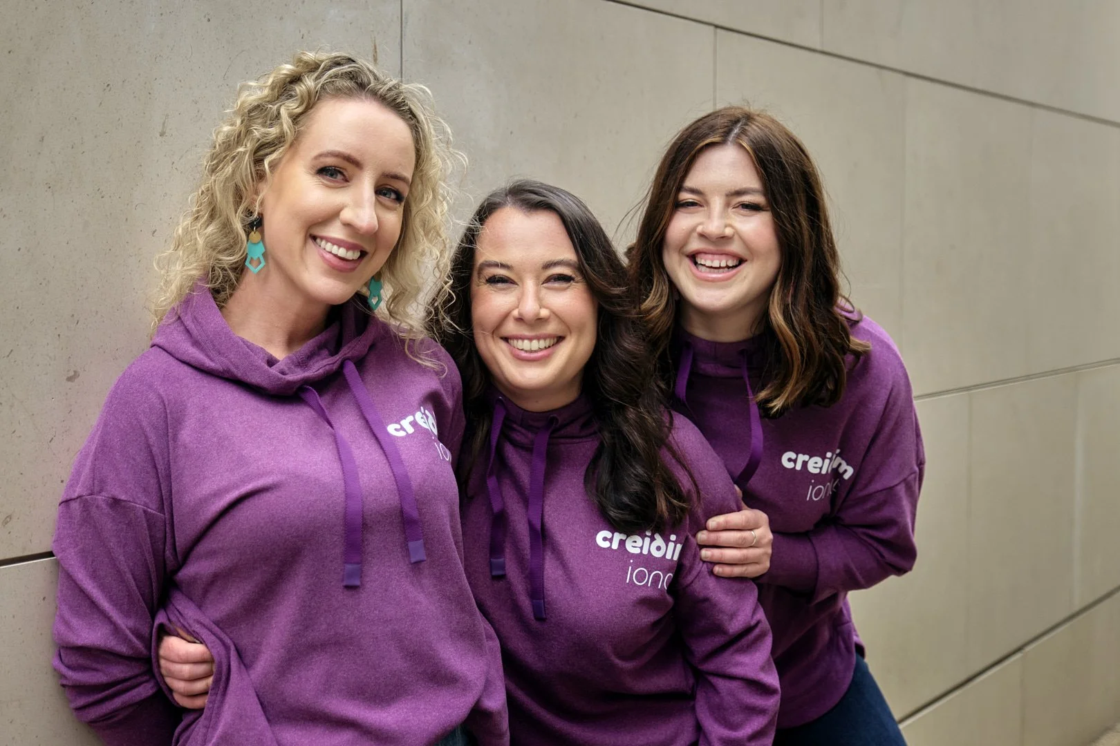 Three women standing close together, smiling, wearing matching purple hoodies with a logo. They are outside against a beige wall.