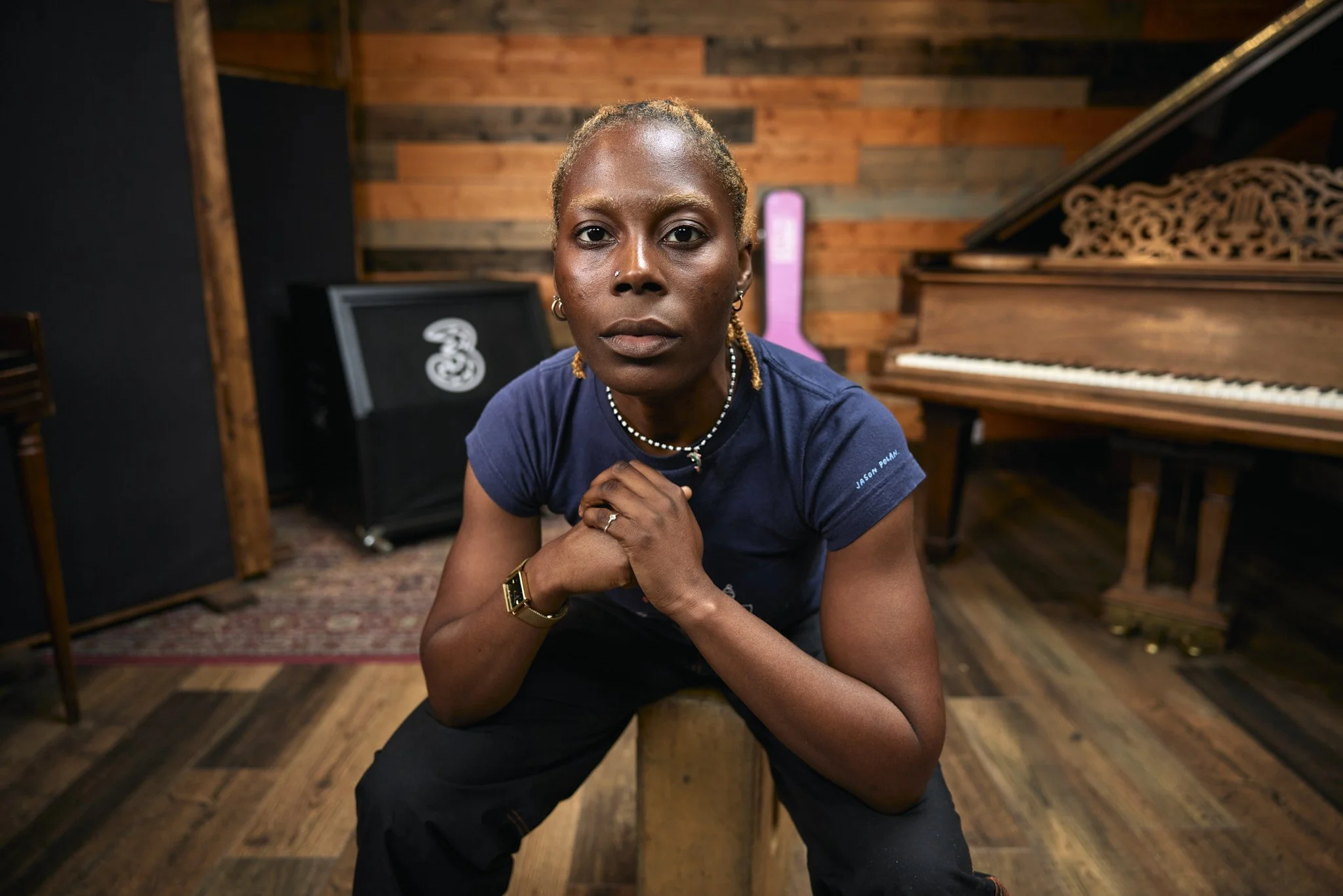 A person with short blond hair and dark skin, wearing a navy t-shirt and jewelry, sitting on a wooden stool in a music studio with a piano and amplifier in the background.