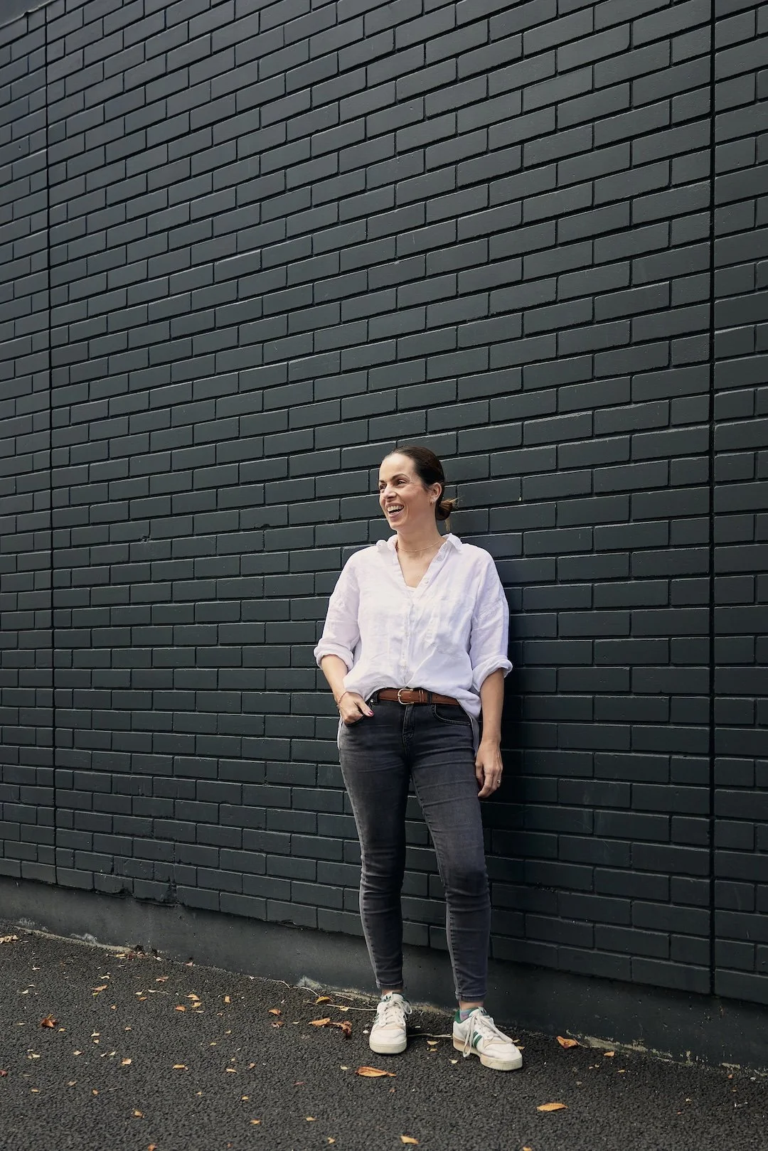 A woman in a white shirt and black jeans laughing and standing against a dark brick wall.
