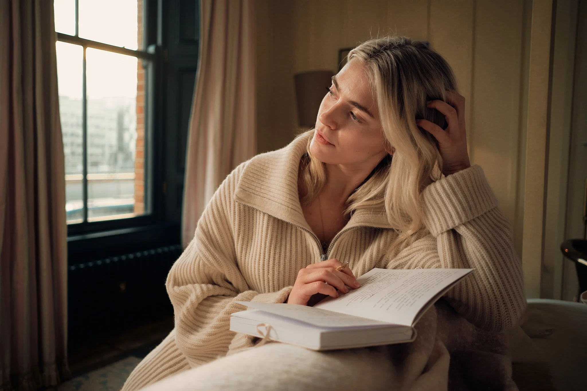 A woman with blonde hair, wearing a beige sweater, is sitting by a window, reading a book, with her head resting on her hand in a cozy room.