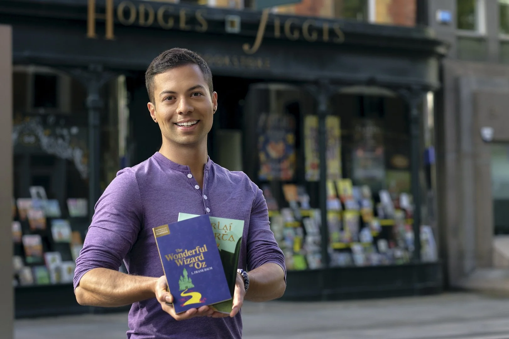 A smiling young man with short dark hair holding three books in front of a bookstore storefront on a city street.