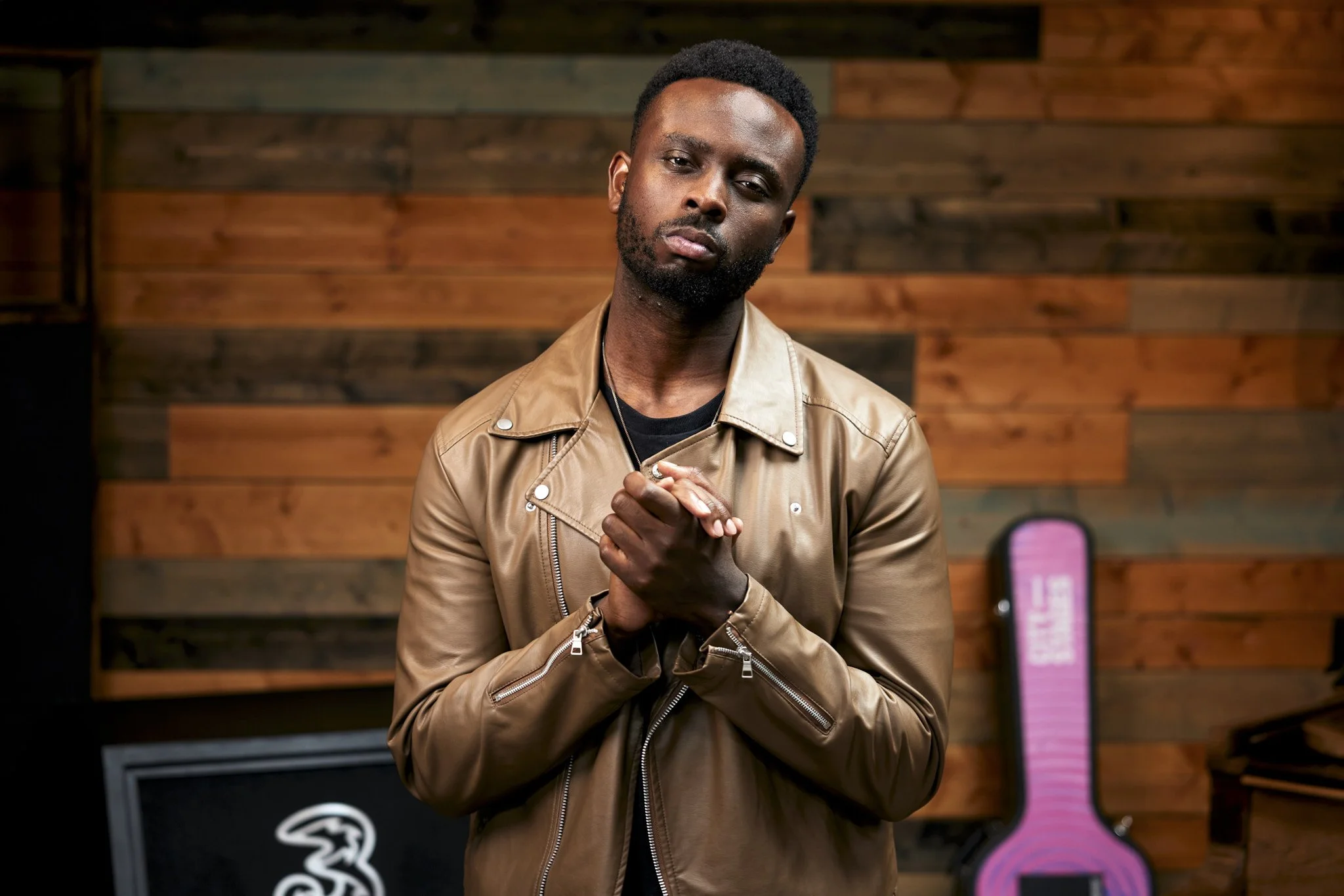 A man with dark skin, short curly hair, and a beard, wearing a tan leather jacket and holding his hands together, stands in front of a wooden-paneled wall with a heart-shaped pink object in the background.