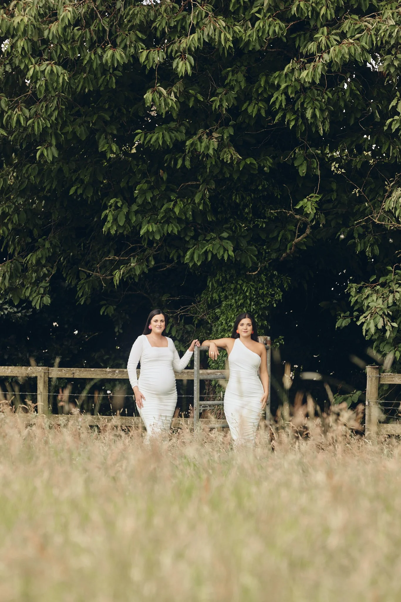 Two women standing in a grassy field in front of a large leafy tree, dressed in white dresses, one with a pregnant belly.