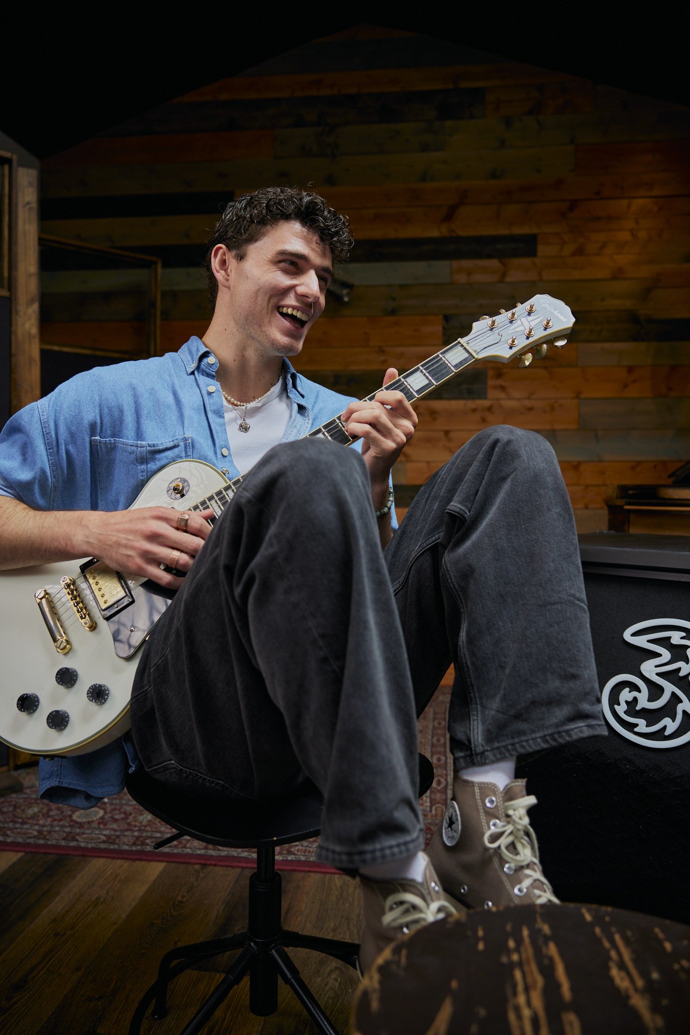 Young man sitting on a chair, playing an electric guitar, smiling, in a music studio with wooden walls.