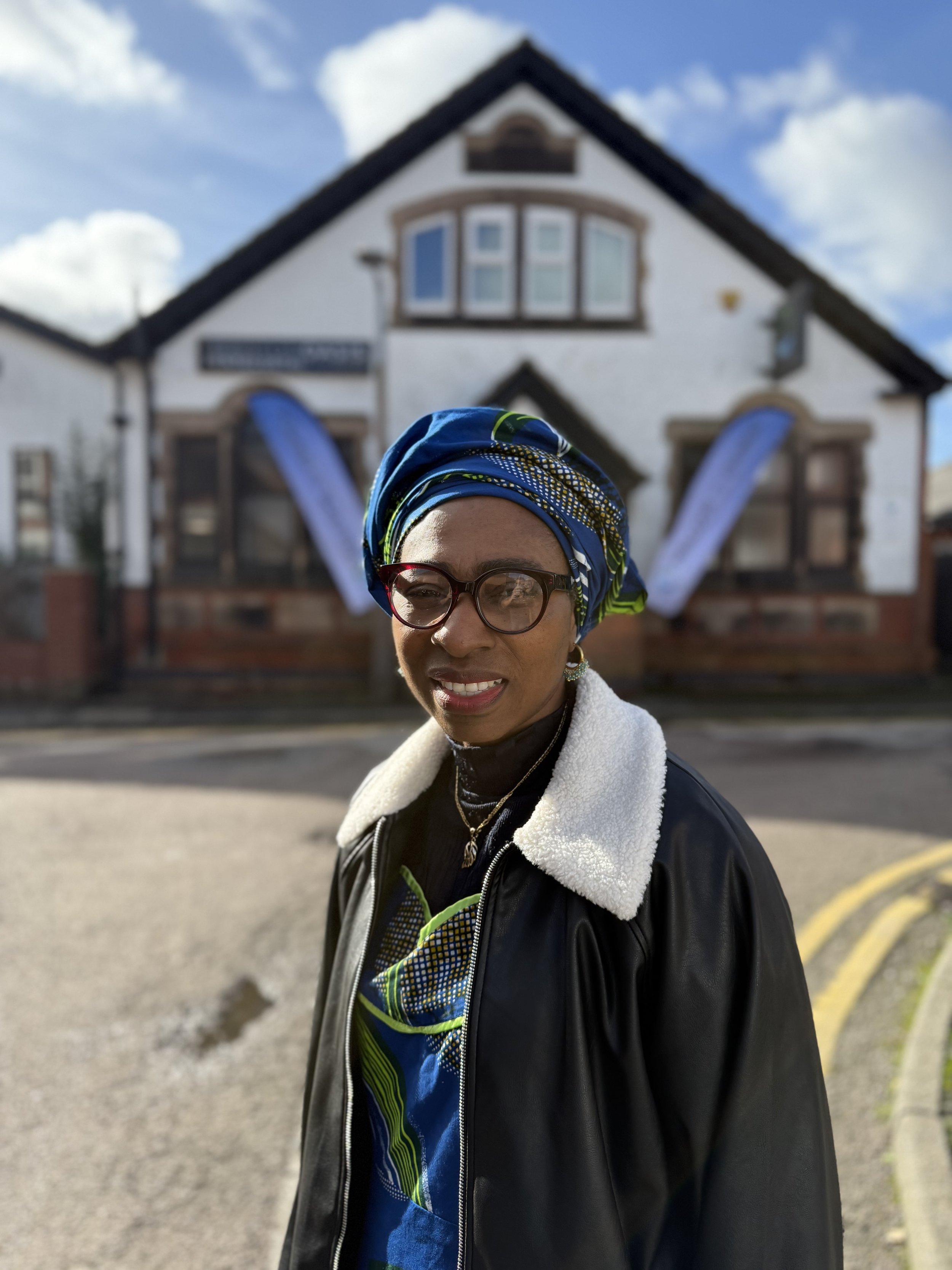 A woman with glasses and a colorful headwrap standing outside in front of a house with a sign and two banners, blue sky with clouds in the background.