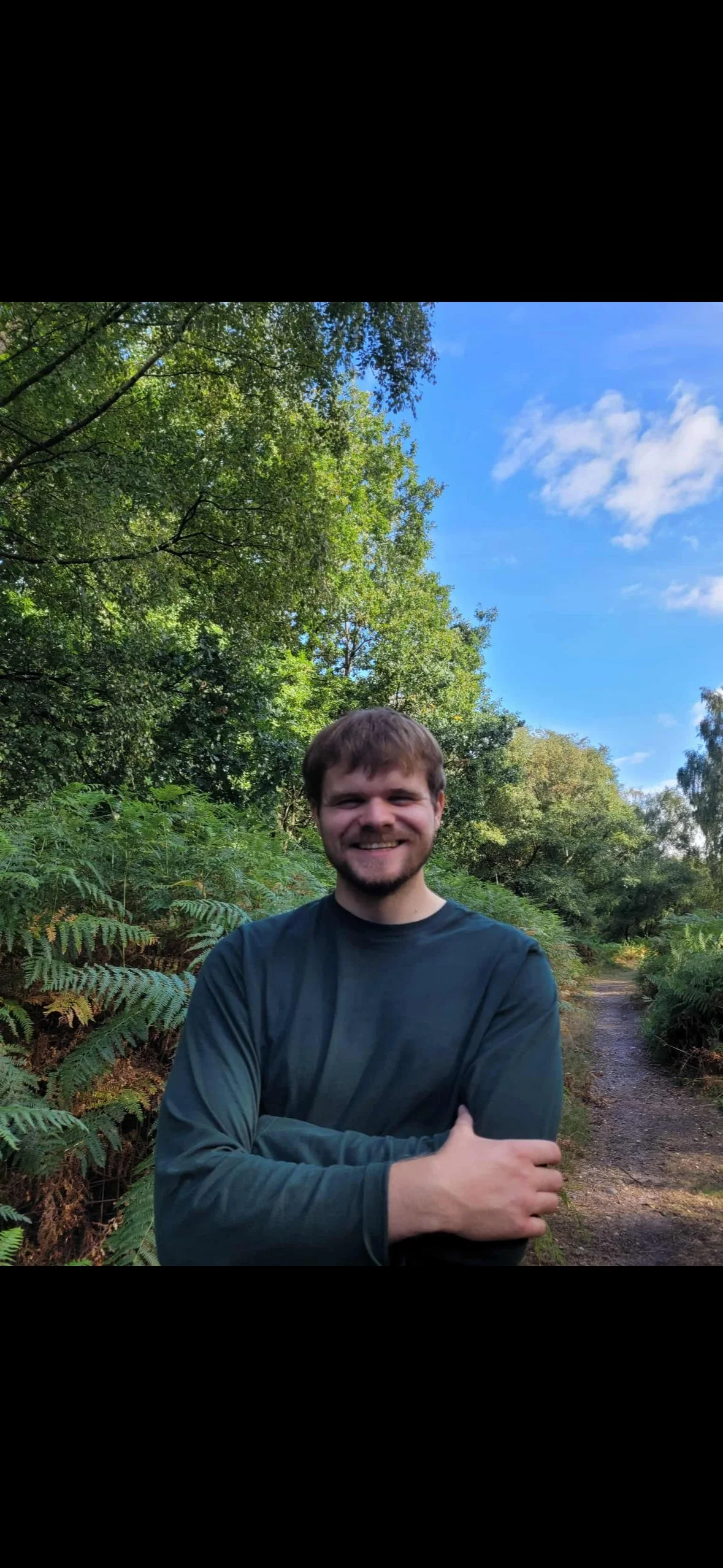 A young man smiling outdoors in a green forest, standing on a dirt path with lush foliage and a partly cloudy blue sky in the background.