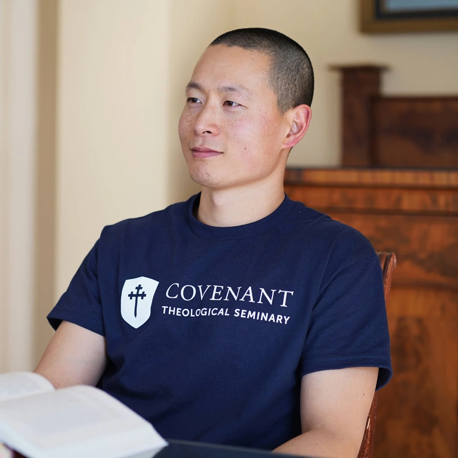 A student sitting at a table, smiling, wearing a navy blue t-shirt with the Covenant Seminary logo