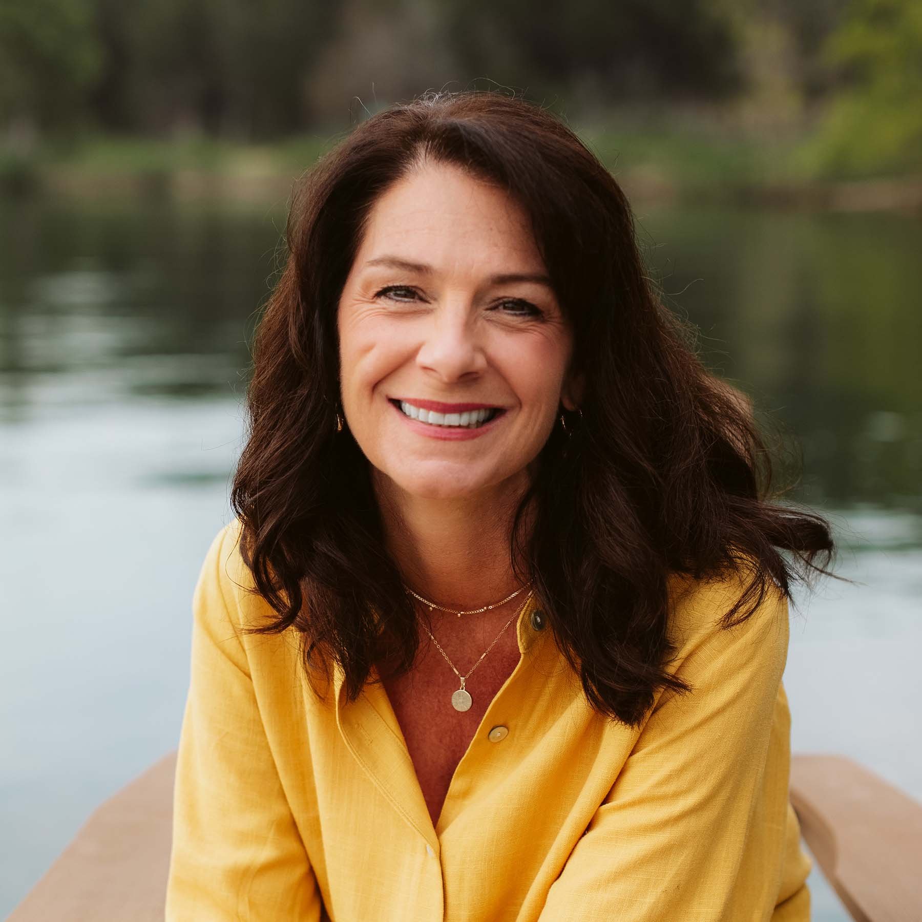 Courtney Doctor smiling outdoors near a body of water, wearing a yellow shirt and necklaces.