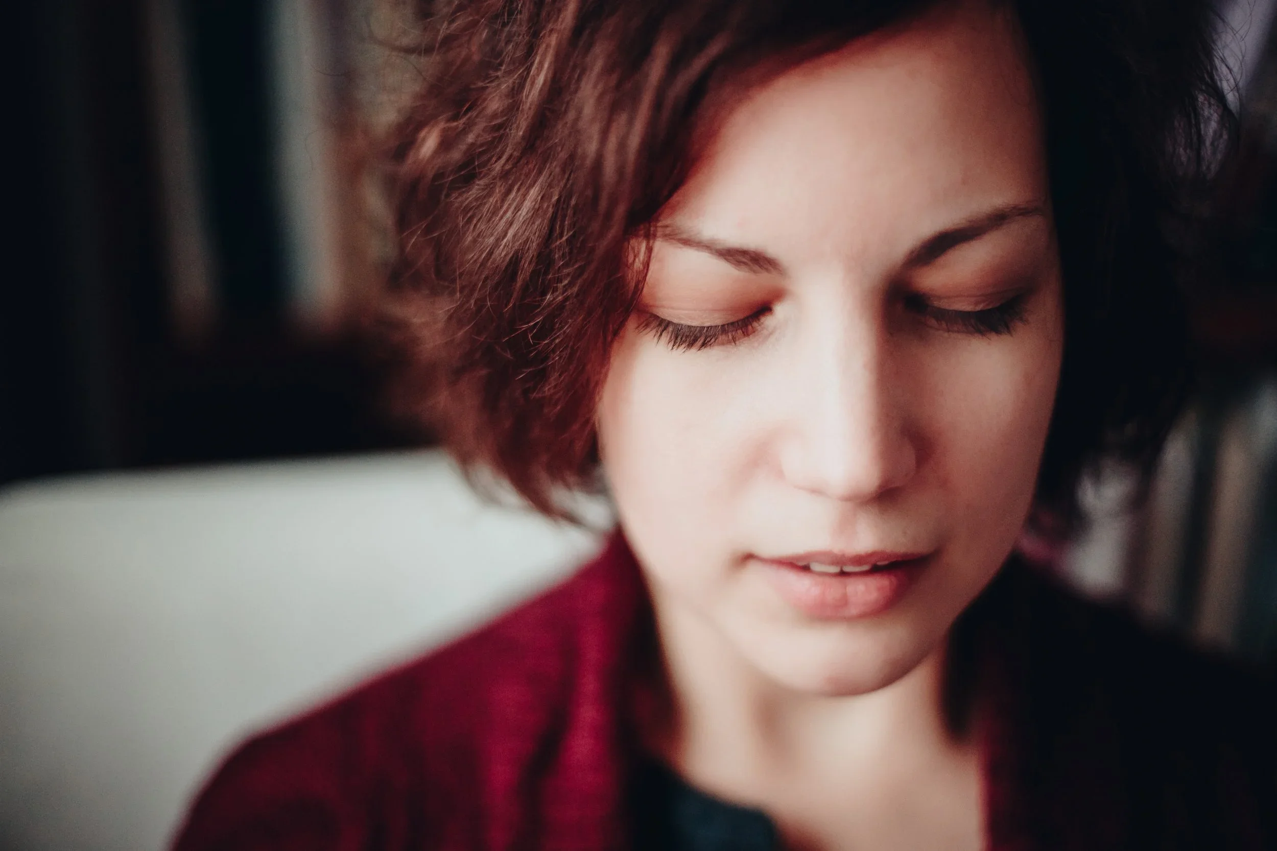 Close-up of a woman with short, wavy brown hair and closed eyes, wearing a deep red garment.