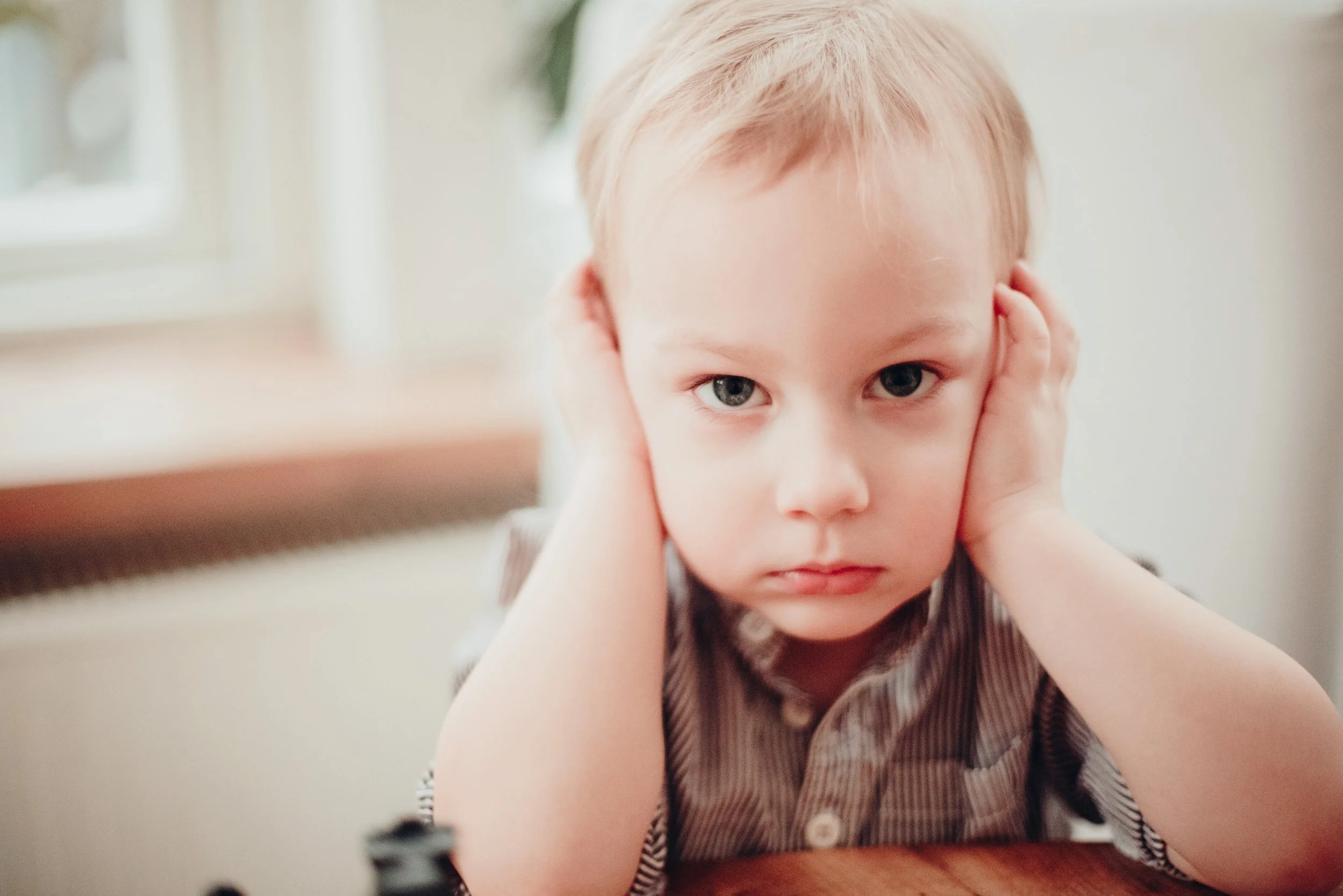A young boy with light hair and blue eyes covering his ears with his hands, looking directly at the camera with a serious expression.