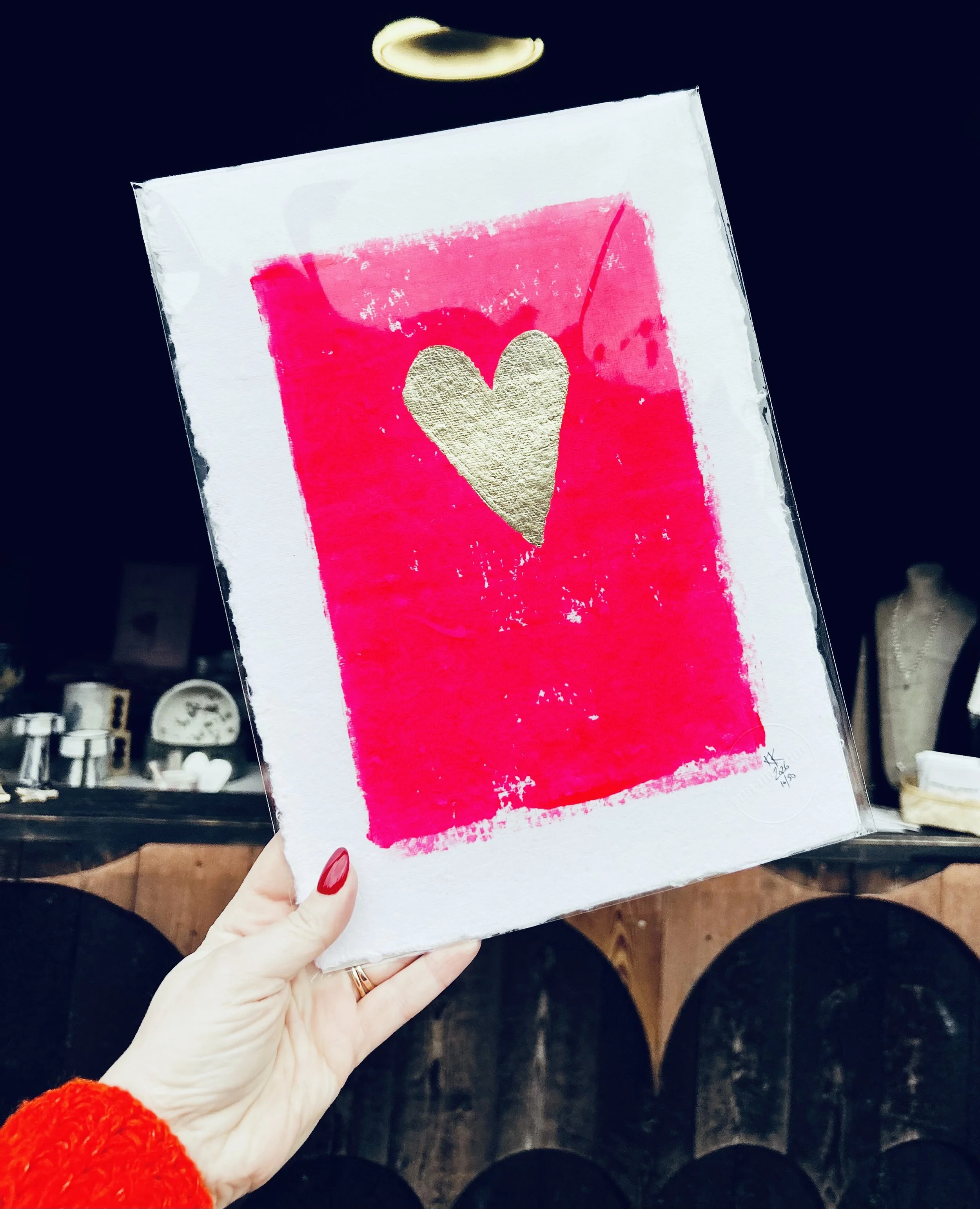 Hand holding a white canvas with a red background and a gold heart in the center.