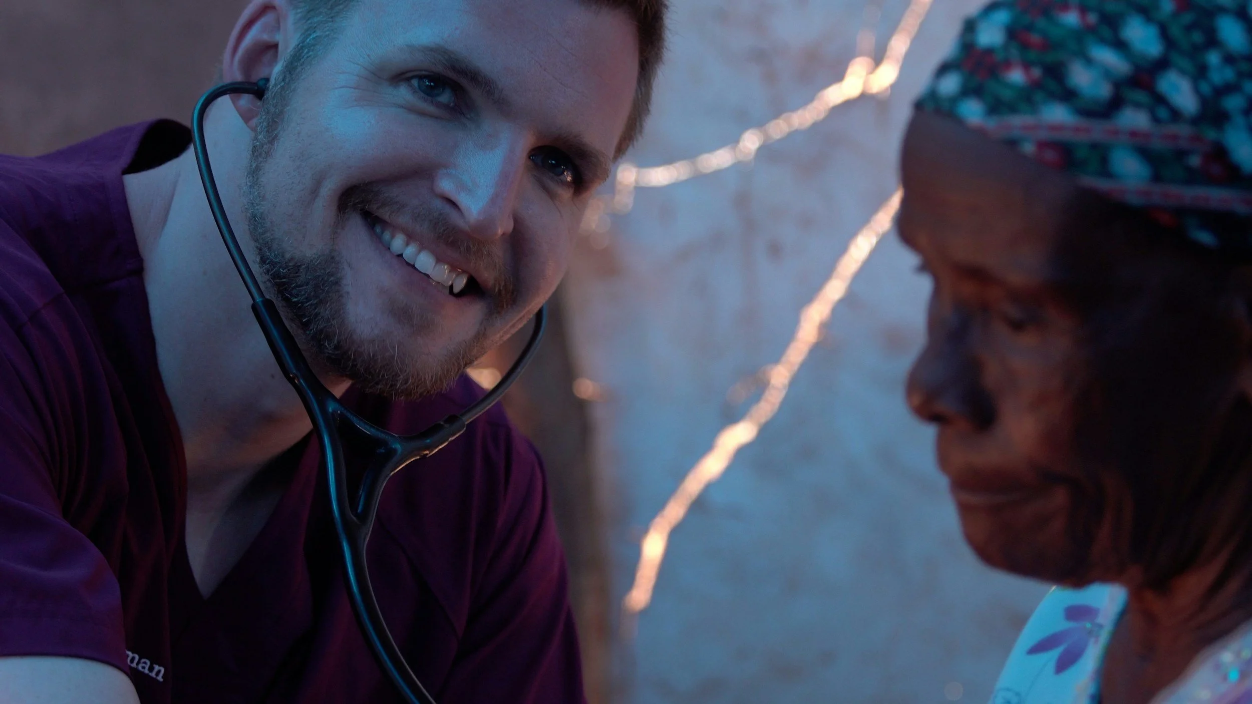 A smiling man with a beard and blue eyes, wearing a maroon shirt and a stethoscope, looking at an elderly woman with dark skin, wearing a colorful headscarf and patterned clothing, in a setting with background lights.