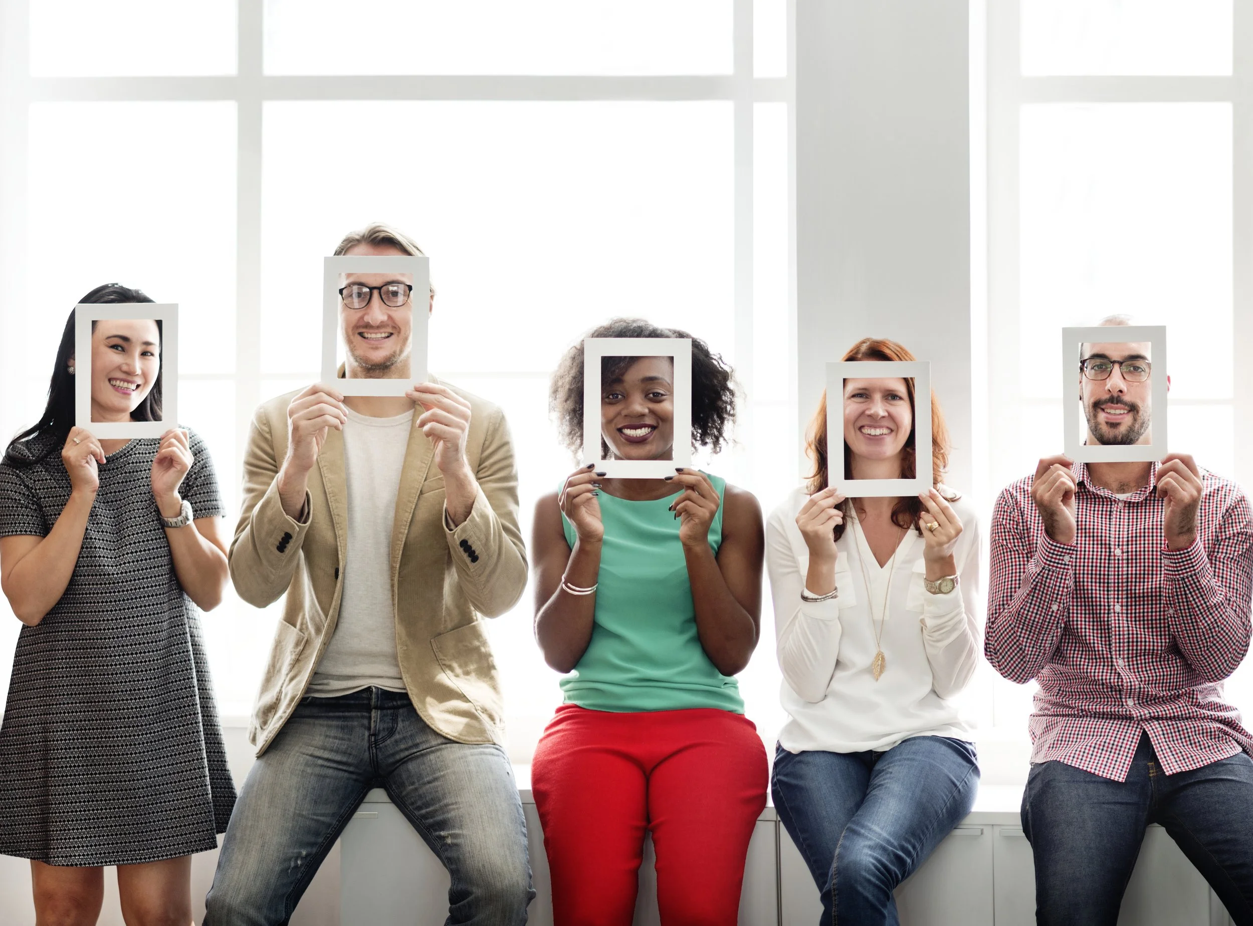 Five diverse people sitting with large windows in the background, each holding a white photo frame around their face, creating a fun group portrait.