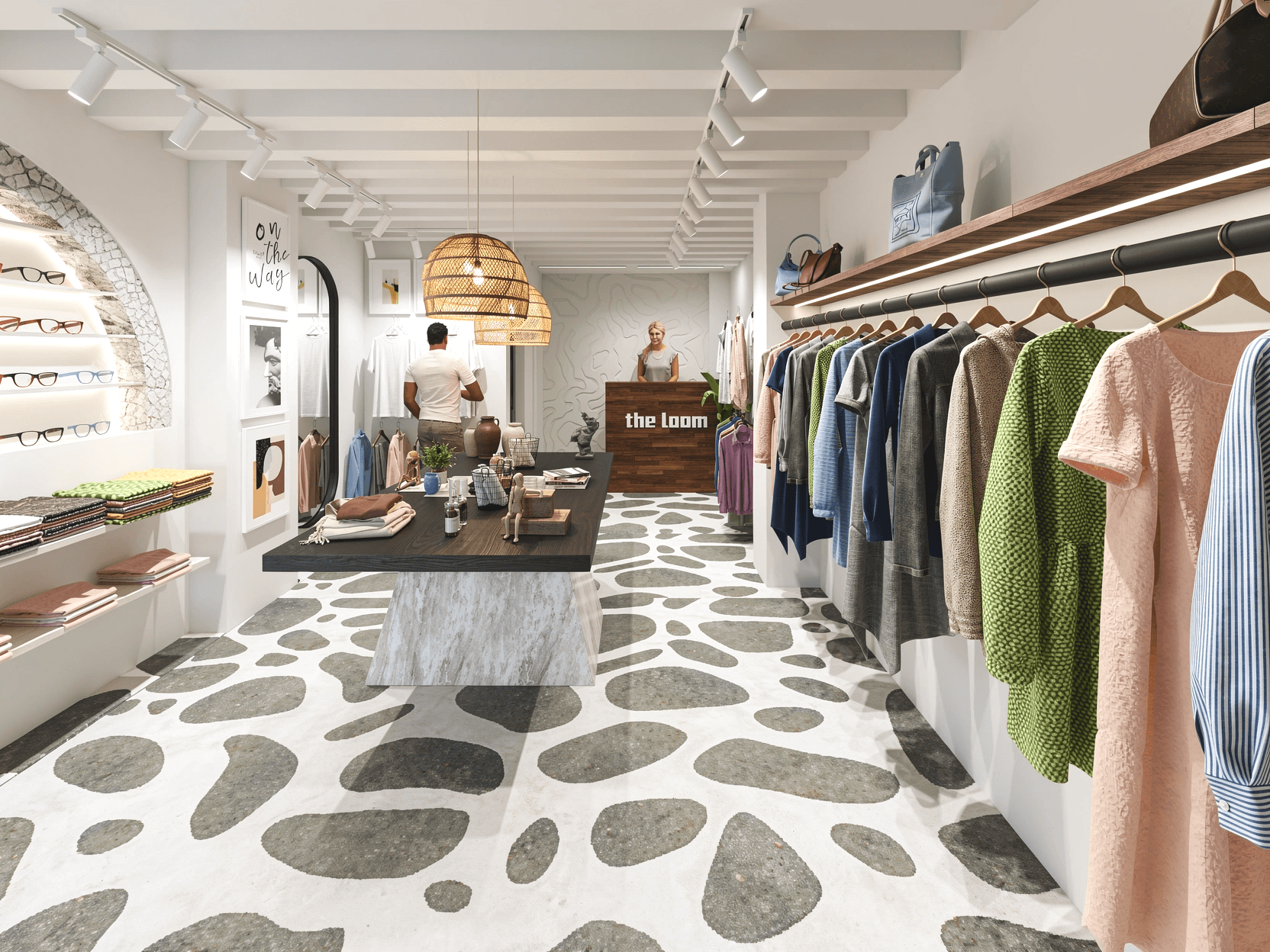 Interior of a clothing store with racks of clothes on the right, a table with accessories in the center, and a counter with a staff member behind it at the back. The store has white walls, patterned floor, and modern decor.