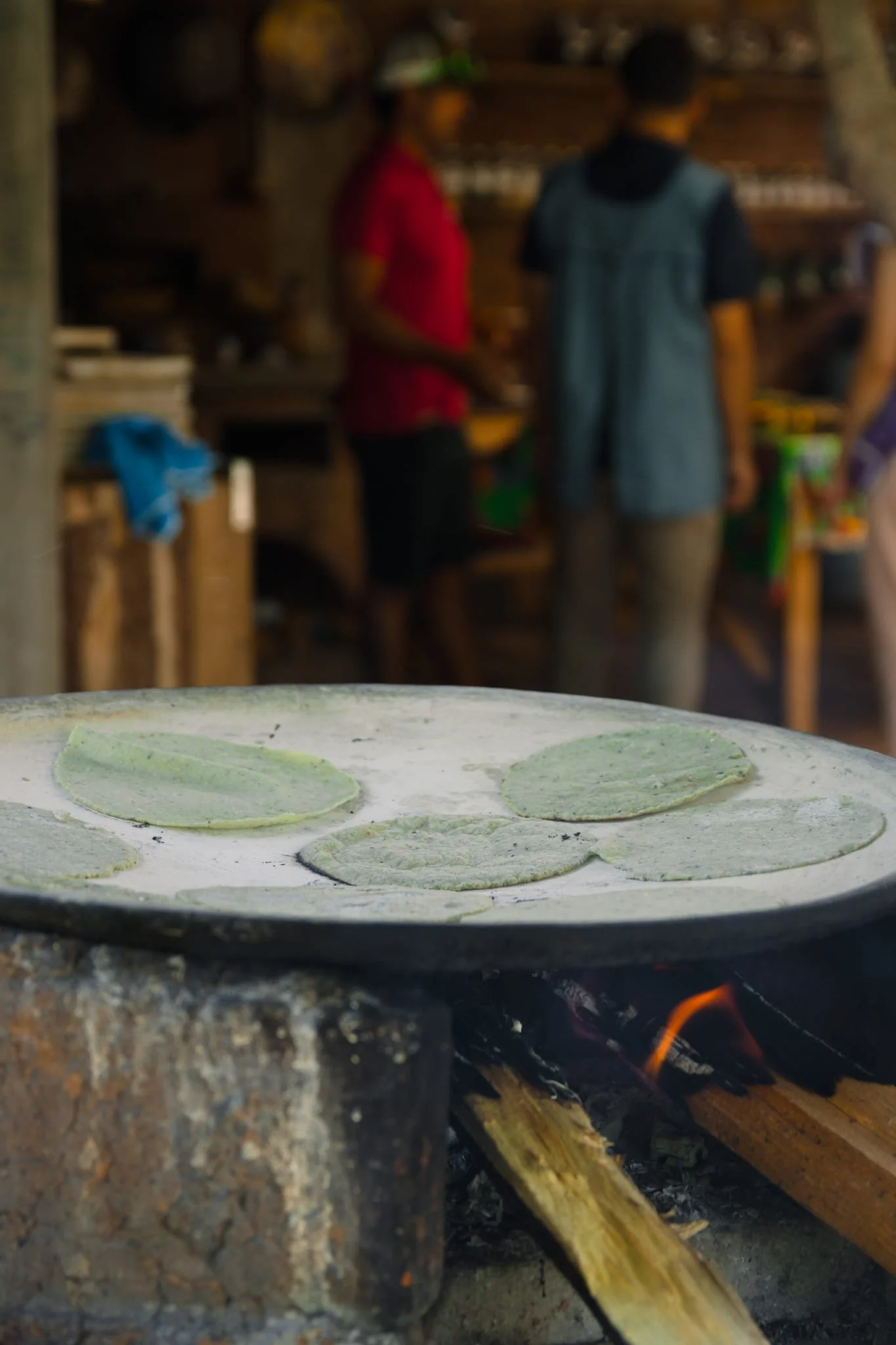 Flat, uncooked green tortillas on a hot griddle over an open fire, with blurred background of two people in a kitchen or outdoor cooking area.
