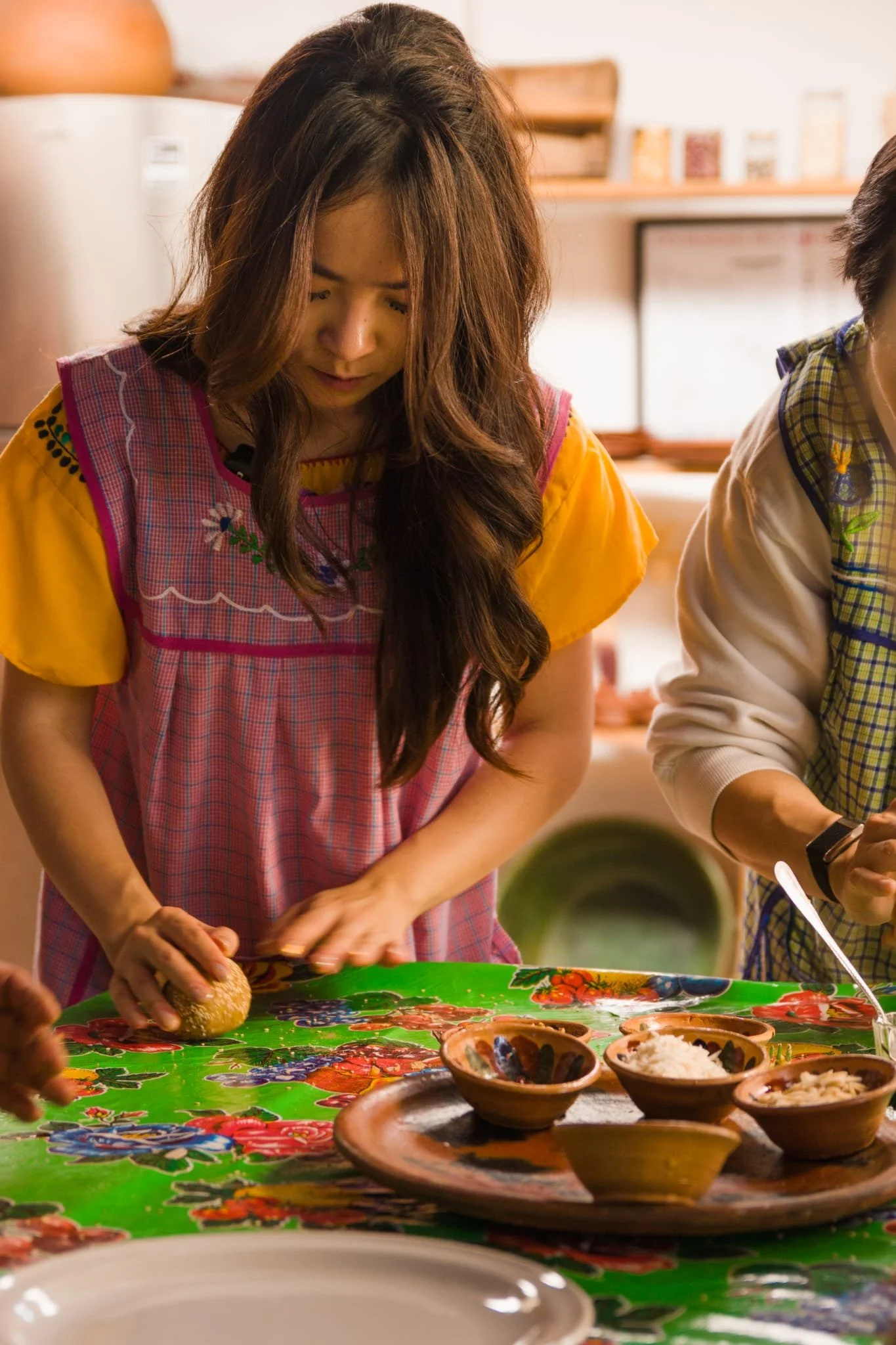 A woman with long brown hair, wearing a colorful dress, is preparing food at a table with a green tablecloth decorated with red and blue patterns. There are multiple small bowls with rice and other ingredients on the table, and she appears focused on