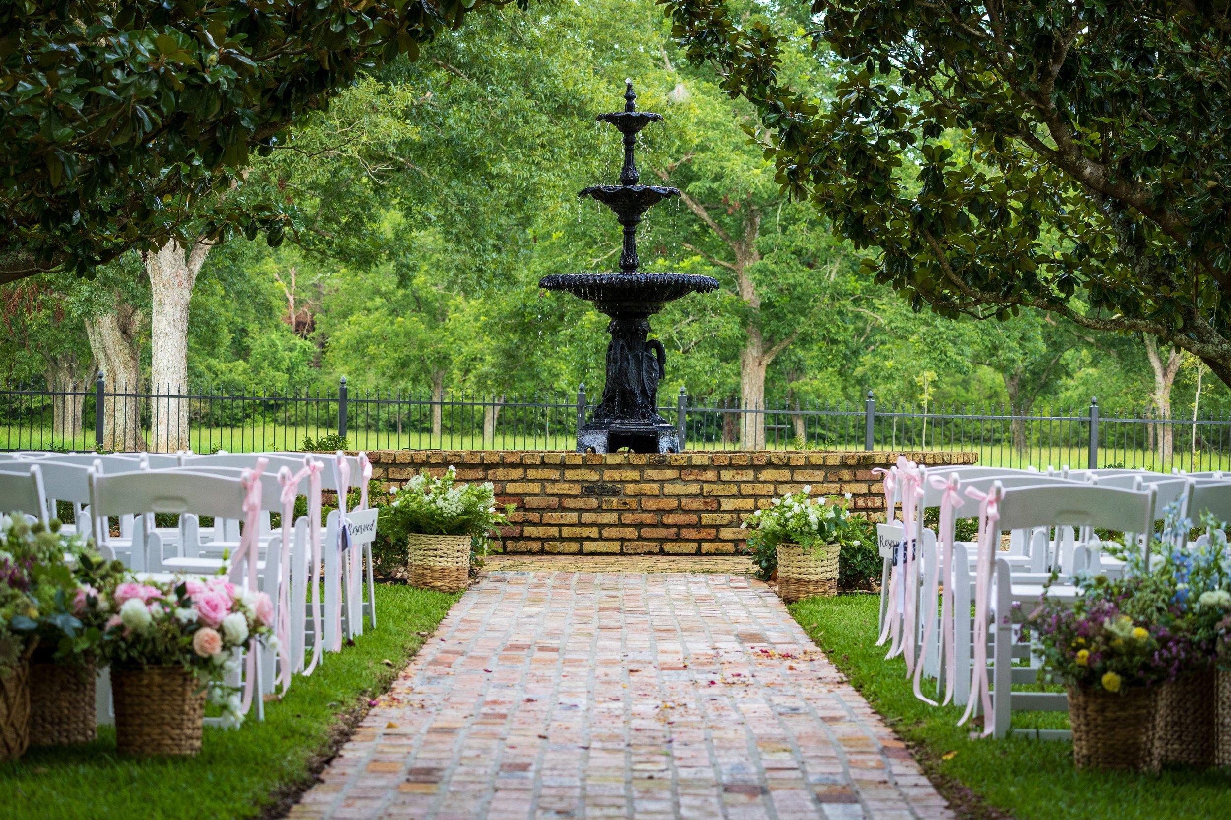 "An outdoor wedding ceremony aisle featuring a brick walkway leading toward a tiered black iron fountain. White folding chairs with pink ribbons line the aisle, accented by woven baskets of pink and white flowers under a canopy of lush green trees."
