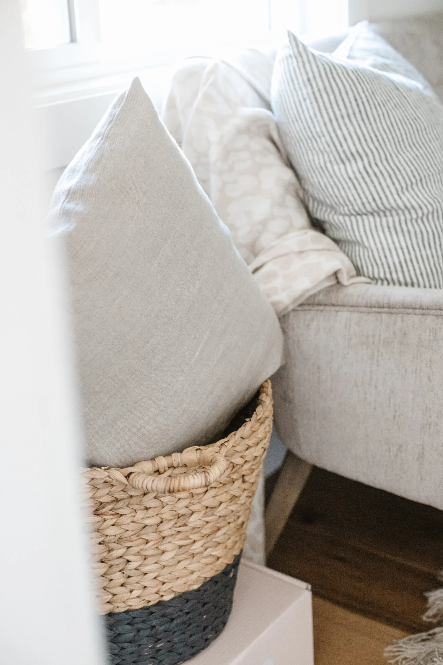 A grey pillow sitting in a light brown wicker basket next to a grey couch that has a light-neutral leopard print throw blanket draped off it.