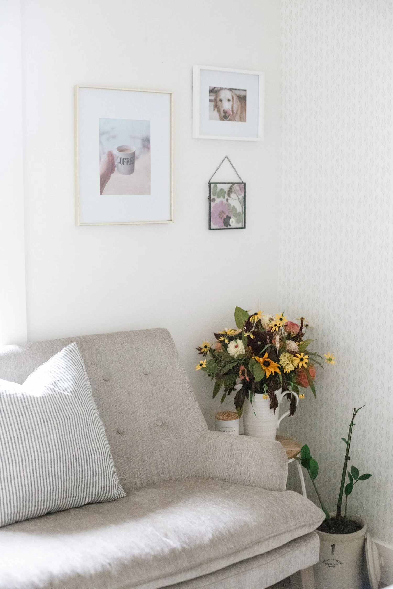 Corner of a space showing a vase of garden flowers sitting on a side table next to a grey couch, with three framed images hanging above on the wall.