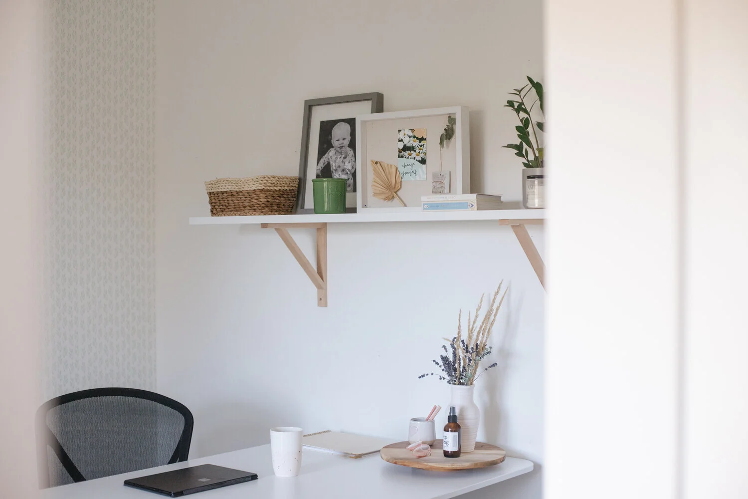 The corner of an office space, with a white shelf hanging above the desk. The shelf has various items on it including framed photos, a candle, a wicker basket & a plant.