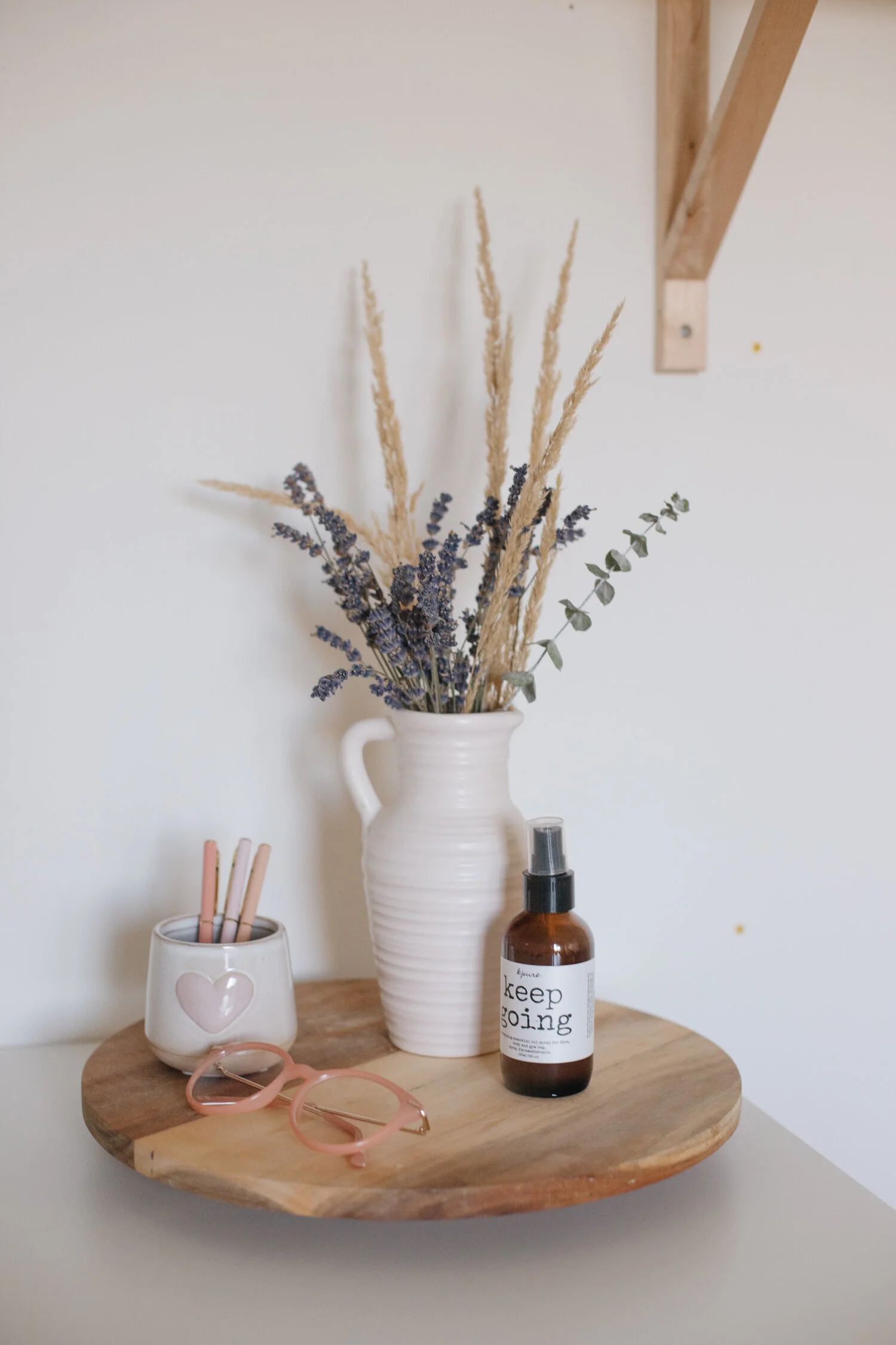 A wooden tray sits on a white desktop holding pink blue-light glasses, a cup filled with pens, a vase with dried florals and a bottle of essential oil spray.