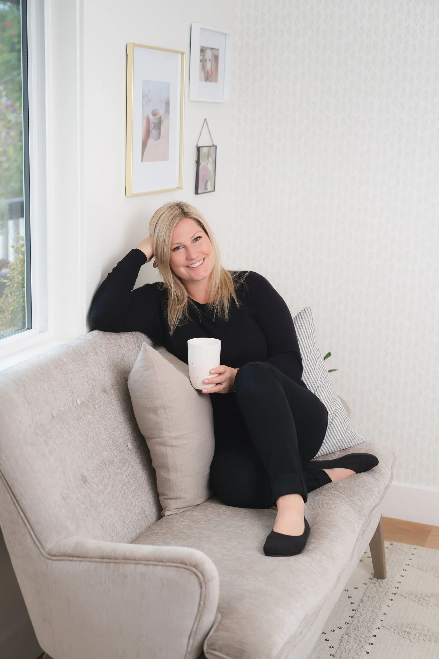 A woman sitting on a couch, holding a cup in one hand, while resting her head in her other hand, smiling at the camera.