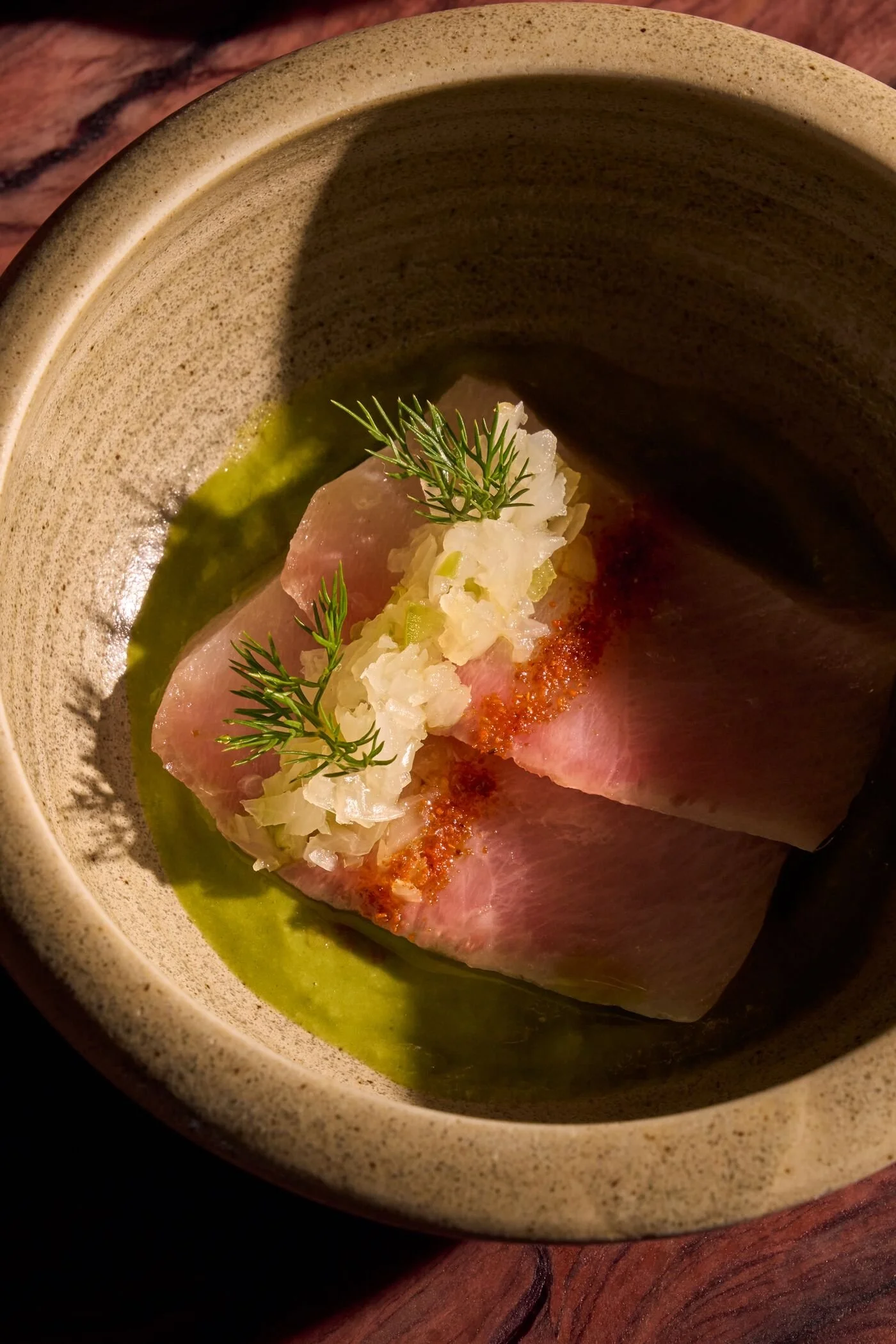 A plate with a slice of raw fish, garnished with fresh herbs, surrounded by green sauce in a beige bowl.
