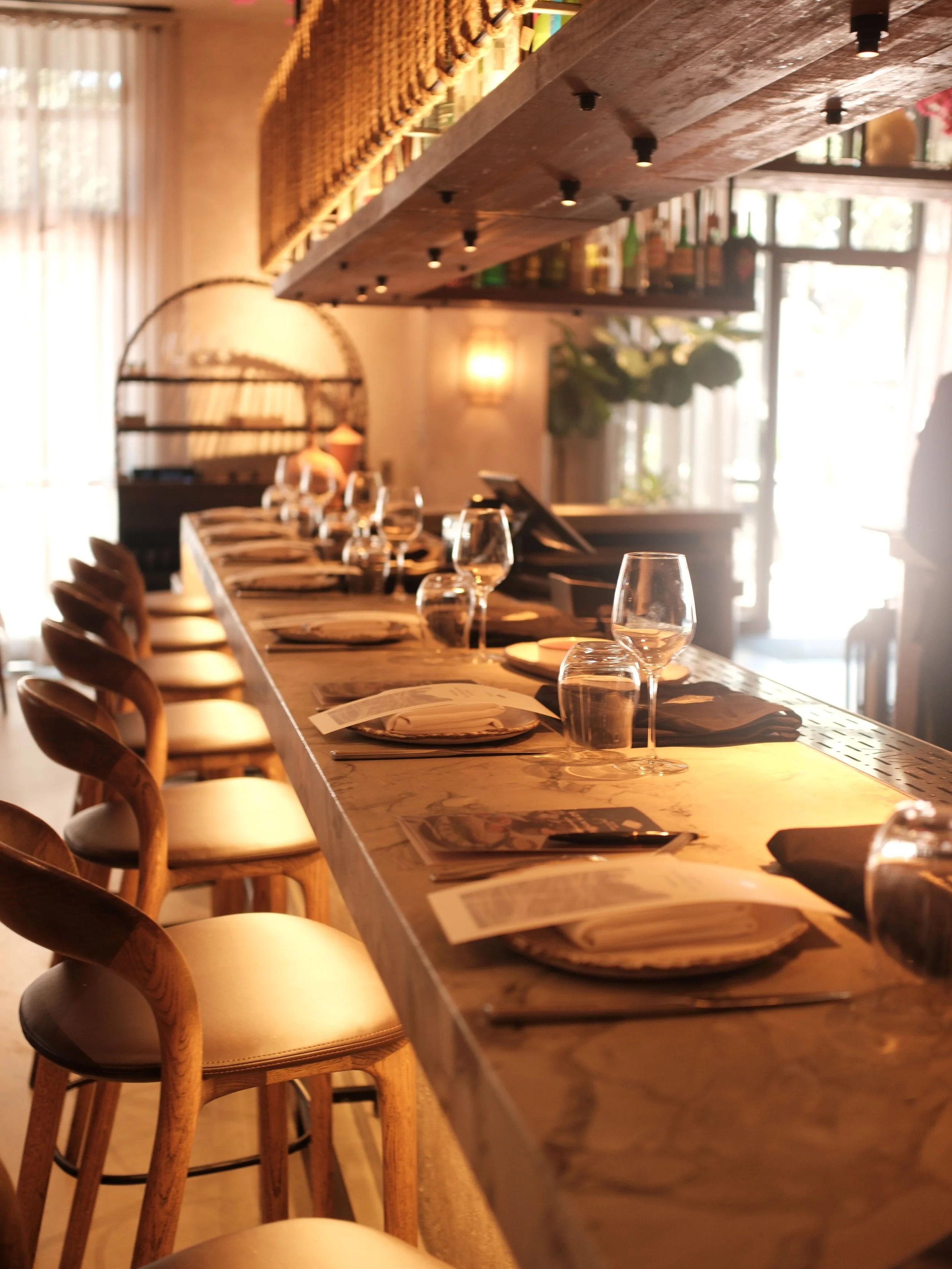A long wooden table set with wine glasses, plates, and napkins in a warmly lit restaurant or cafe with natural light coming from large windows, surrounded by wooden chairs with curved backs.