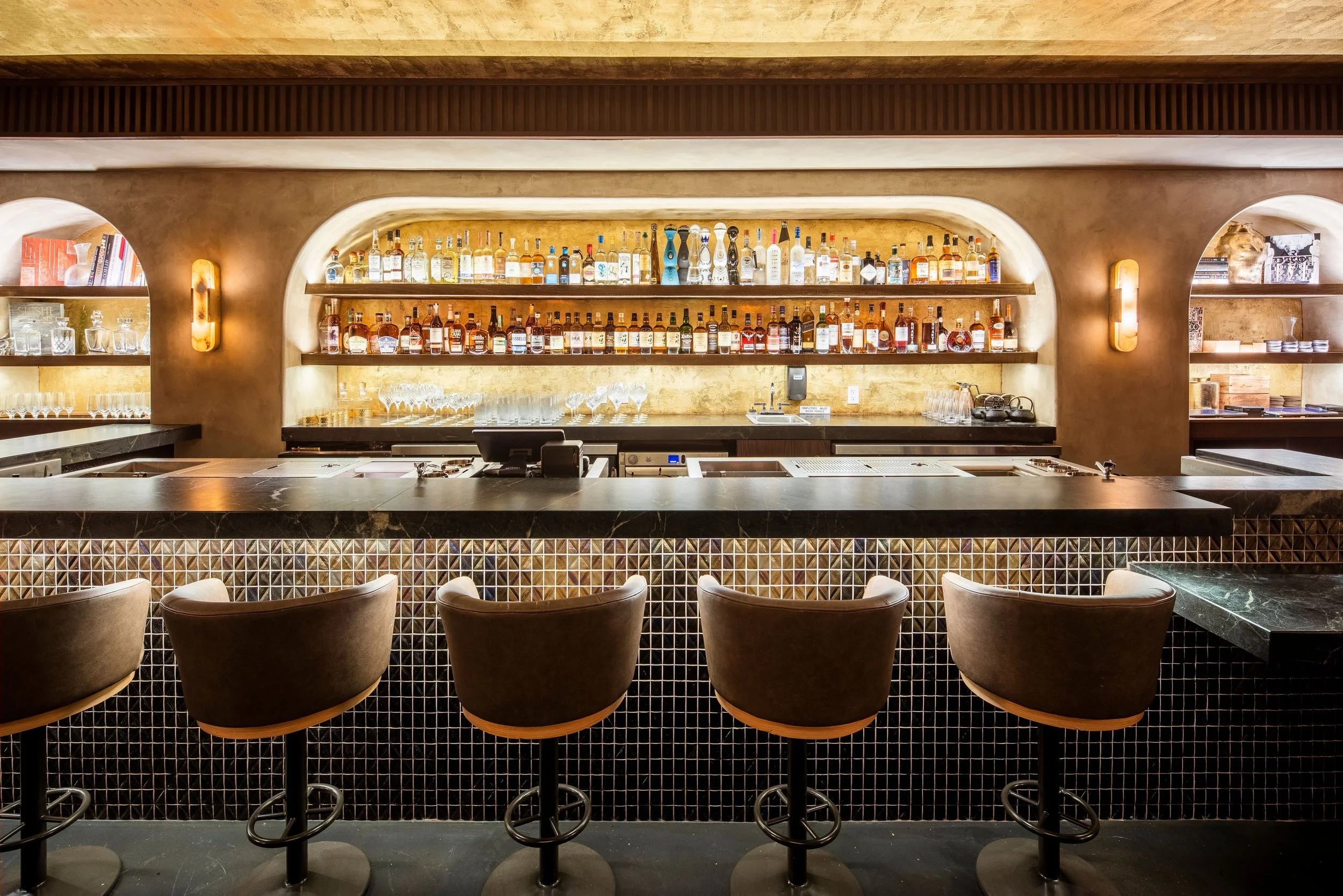 A bar with shelves of various bottles of alcohol, a row of glasses, and bar stools in front of a black marble counter.