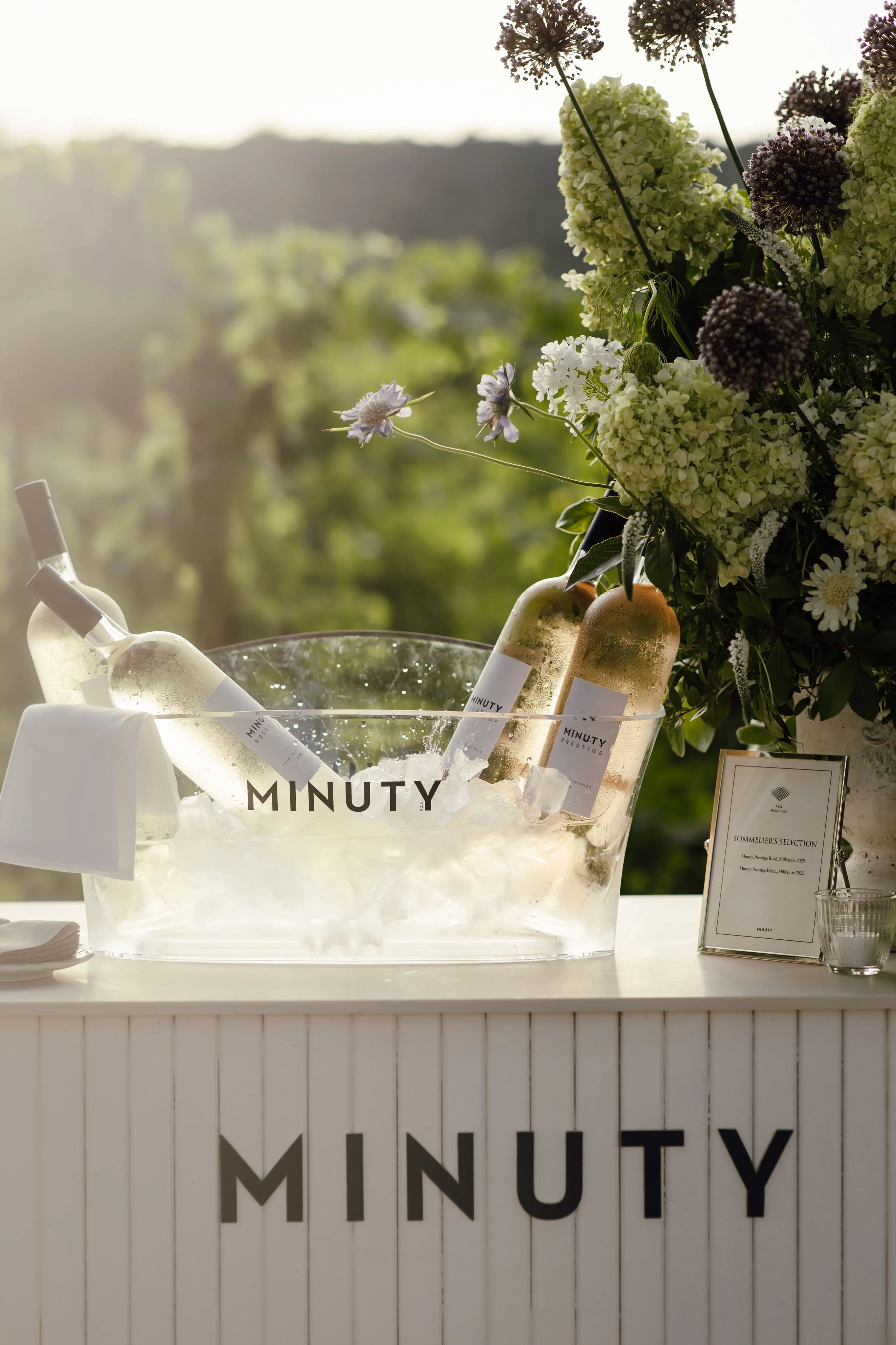 A display of bottled beverages in a clear ice-filled container at an event, with a bouquet of white and purple flowers in the background, and a sign with the brand name 'MINUTY' on the front of the container.
