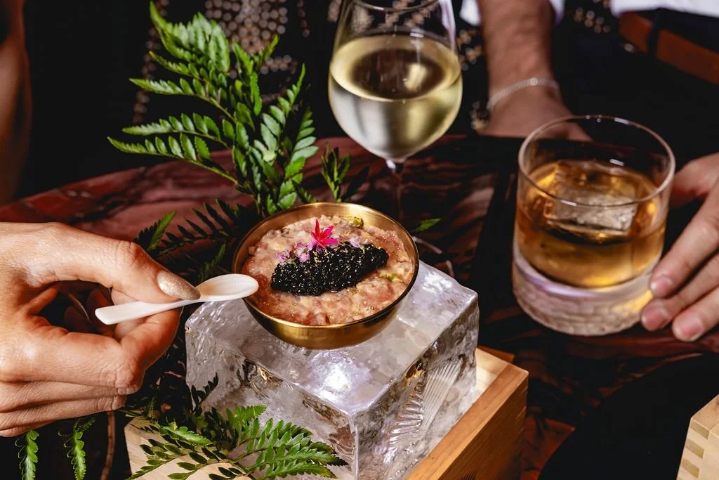 A hand holding a small spoon over a dish topped with black caviar and garnished with a pink flower. The dish is placed on ice on a wooden tray, with green fern leaves nearby. Two drinks are on the table: a glass of white wine and a glass of whiskey with ice.