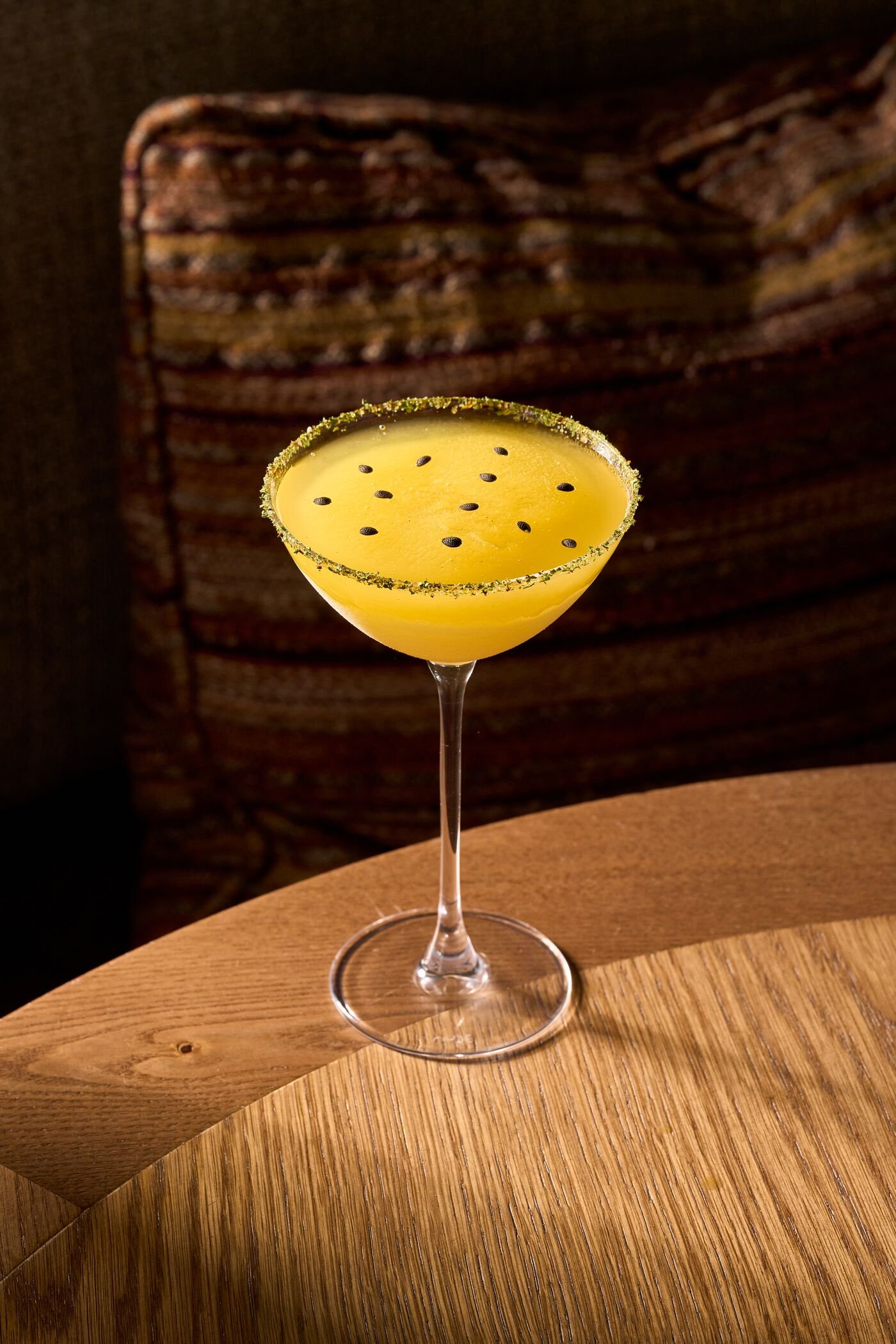 A yellow cocktail with black seeds rimmed with green salt, served in a martini glass, on a wooden table with a textured brown background.