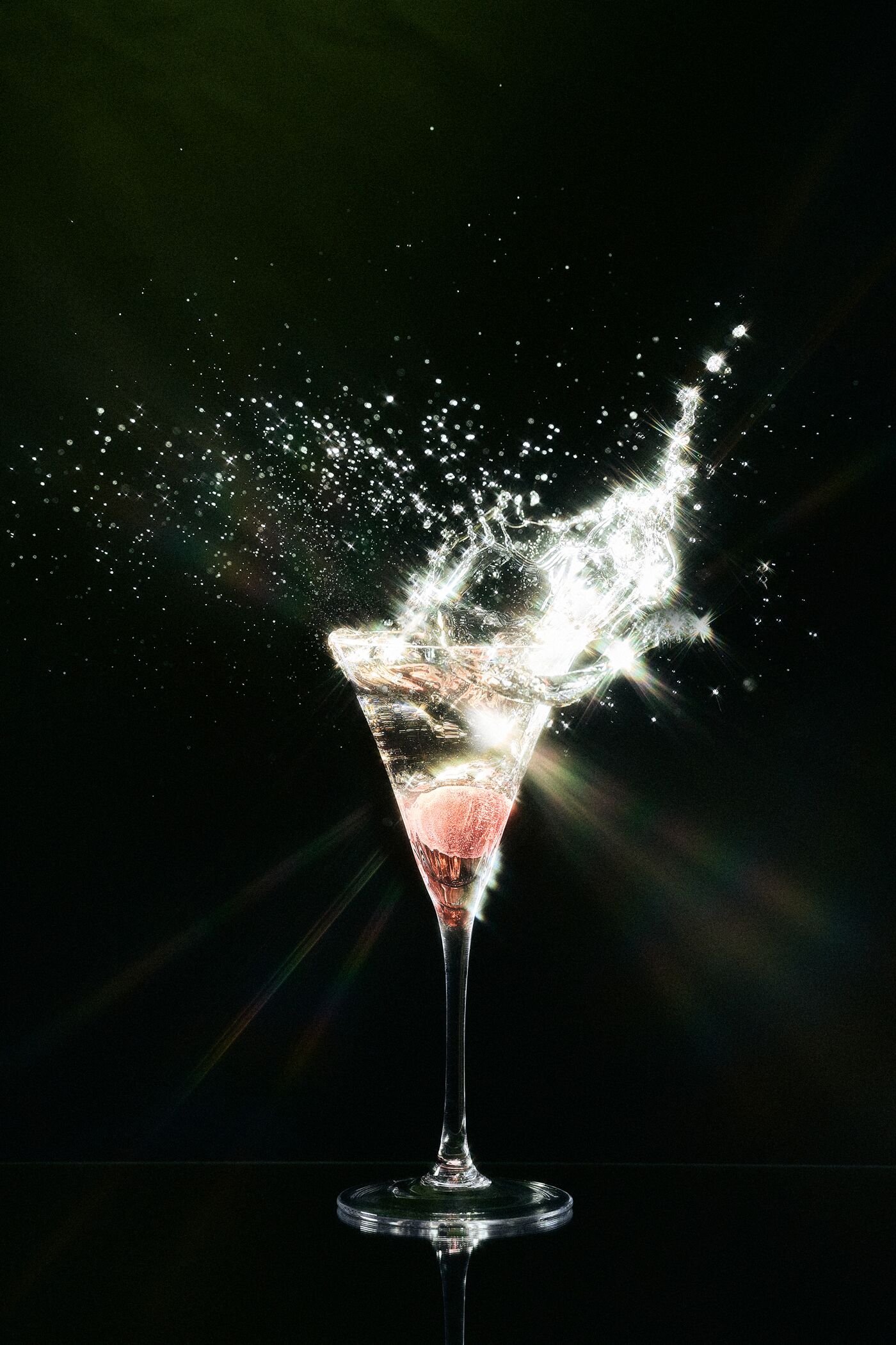 A tall, elegant champagne flute filled with pink liquid, with water or champagne splashing out of the top, creating a burst of sparkling droplets against a dark background.