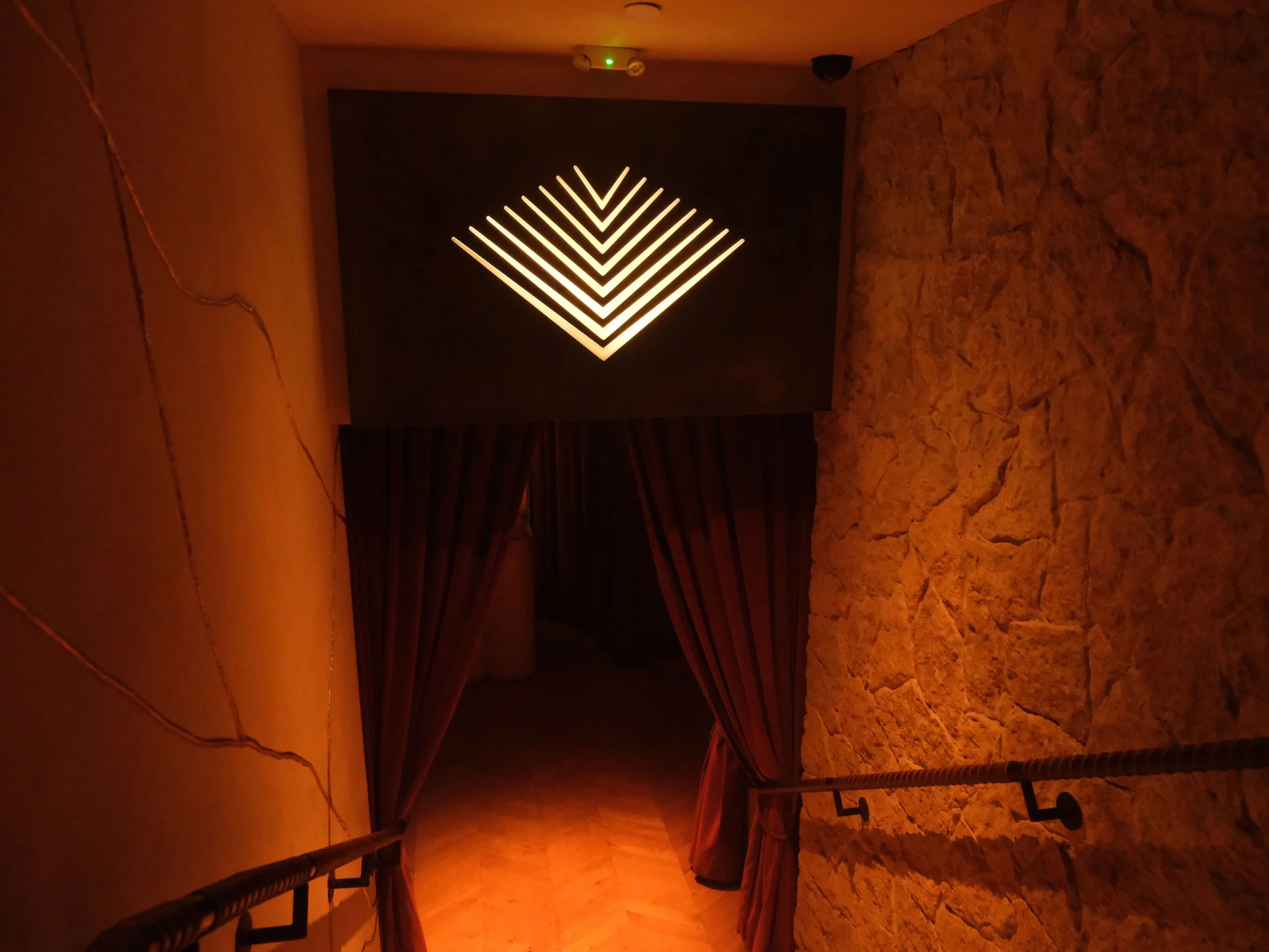 Interior view of a dimly lit room with warm lighting and exposed brick walls. A modern light fixture with multiple rows of downward-facing lights is mounted on a dark wall. The image is rotated 90 degrees clockwise.