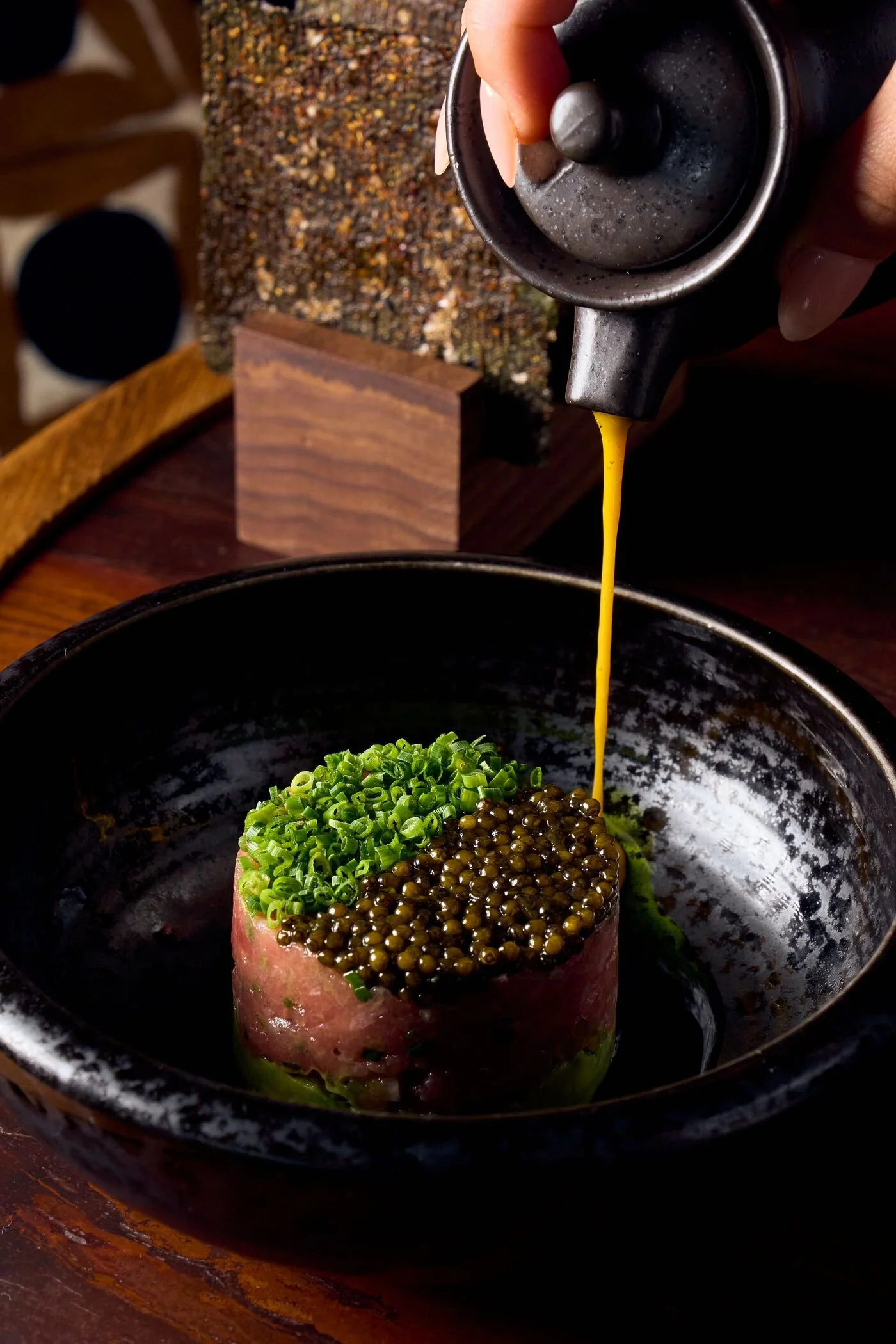 A person pouring a yellow liquid, likely sauce or broth, onto a dish with green onions and caviar in a black bowl on a wooden table.