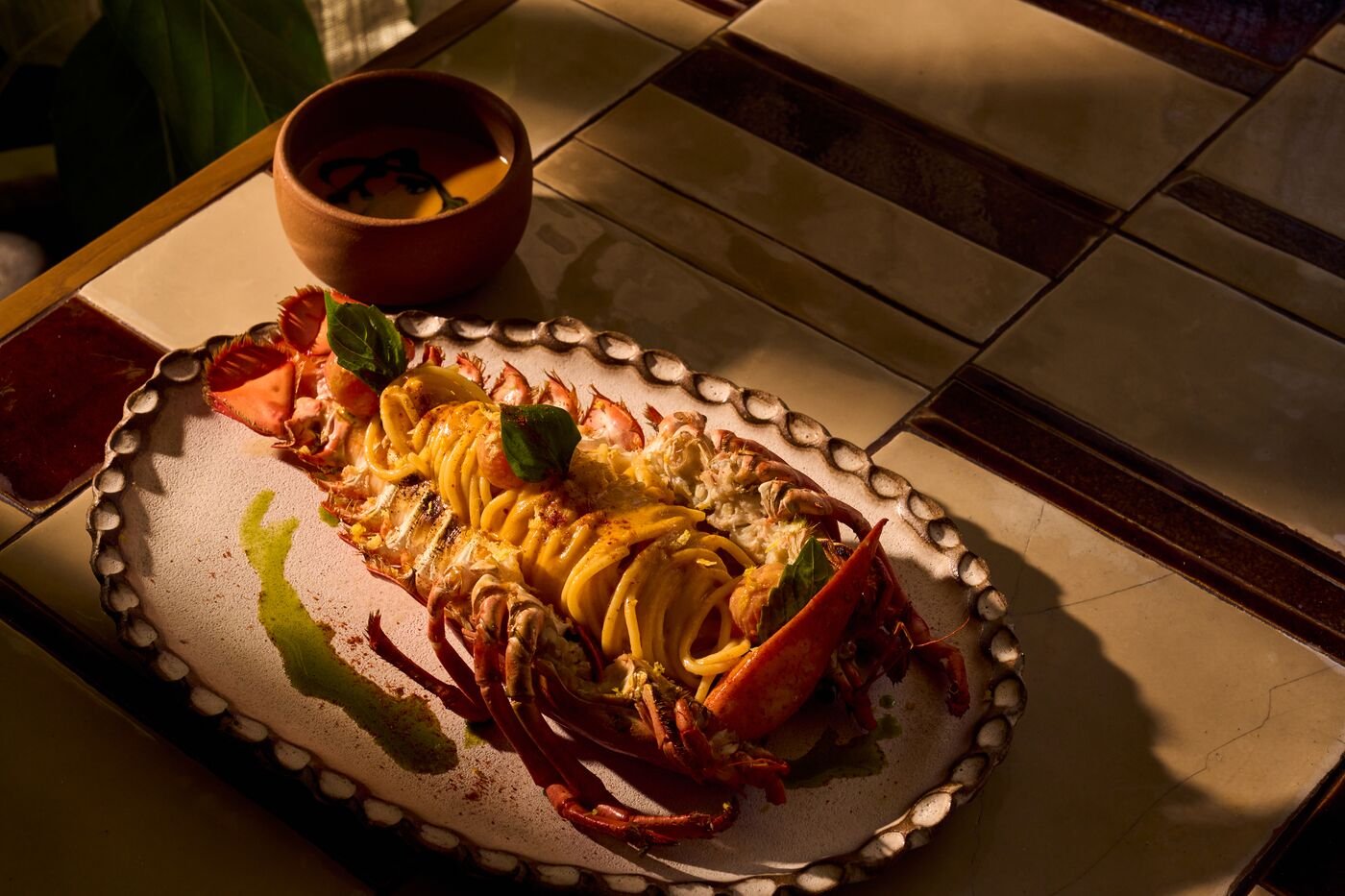 Lobster tail served with spaghetti and garnished with herbs on a decorative oval plate, with a small bowl of sauce in the background.