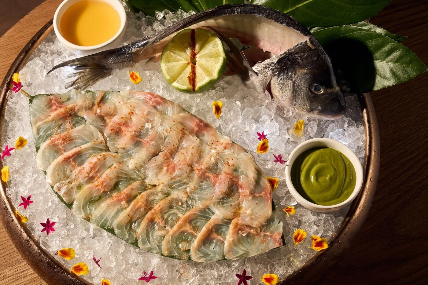 A seafood platter with sliced fish, a whole fish, lime, and sauces on ice, decorated with flower petals.