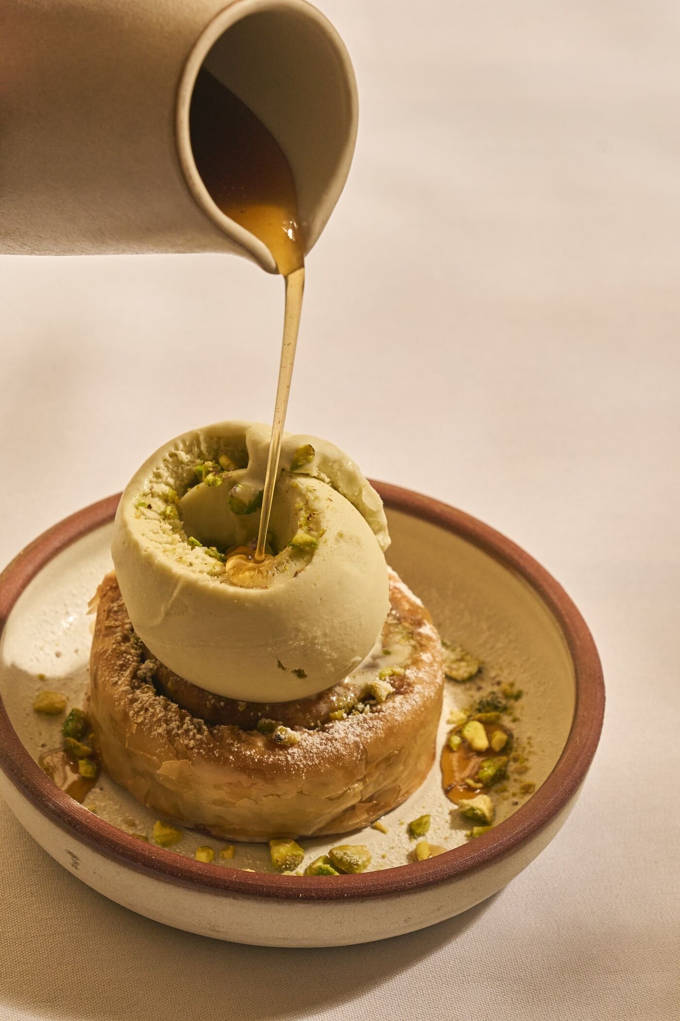 A scoop of vanilla ice cream topped with chopped pistachios, with syrup being poured over it from a cup. The bowl is on a white surface.