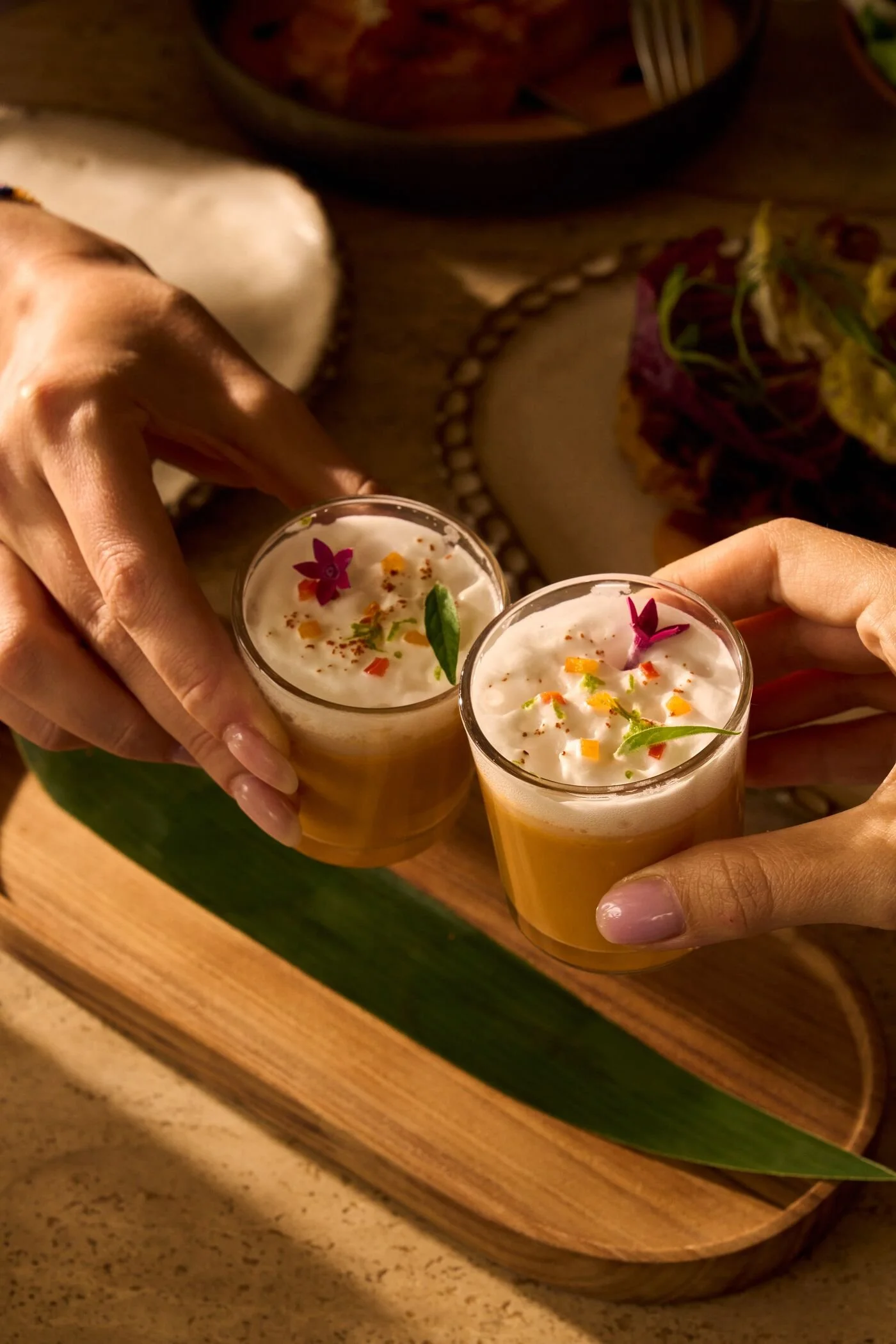 Two glasses of mango lassi with topping of edible flowers and herbs, held by three people above a wooden tray with a green leaf.