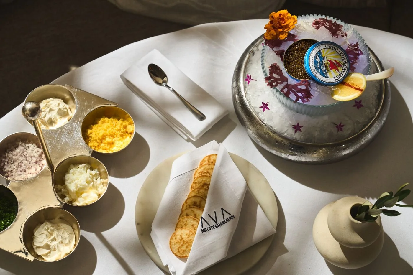 A table with various condiments, a wrapped breadstick, and a bowl of caviar with lemon, flowers, and ice