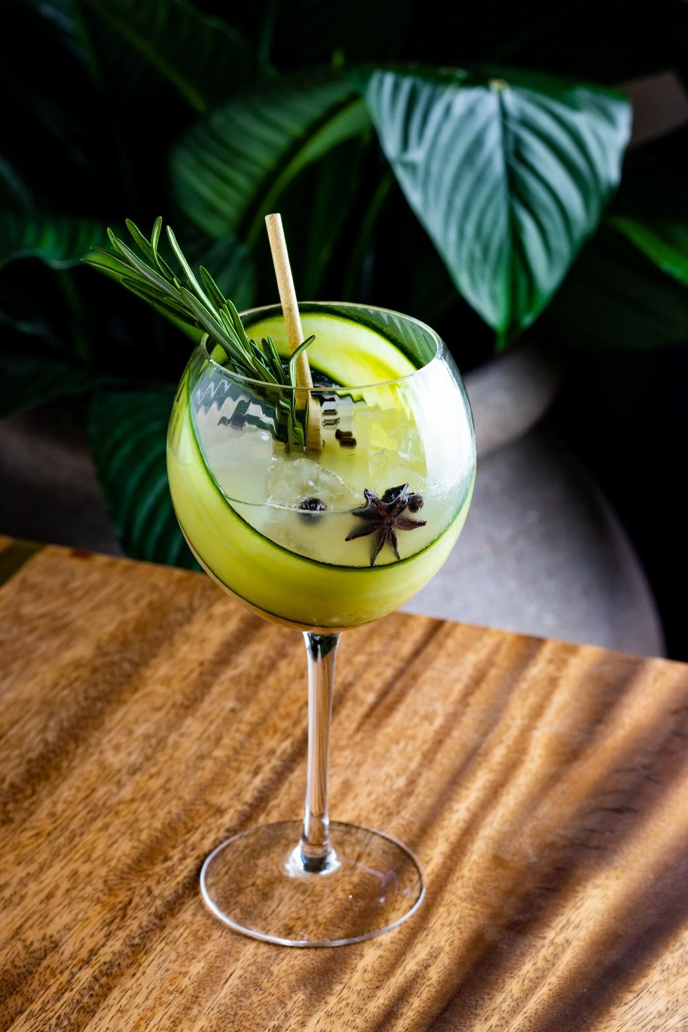 A cocktail in a large wine glass garnished with a cucumber slice, a sprig of rosemary, star anise, and a cinnamon stick, placed on a wooden surface with green tropical leaves in the background.
