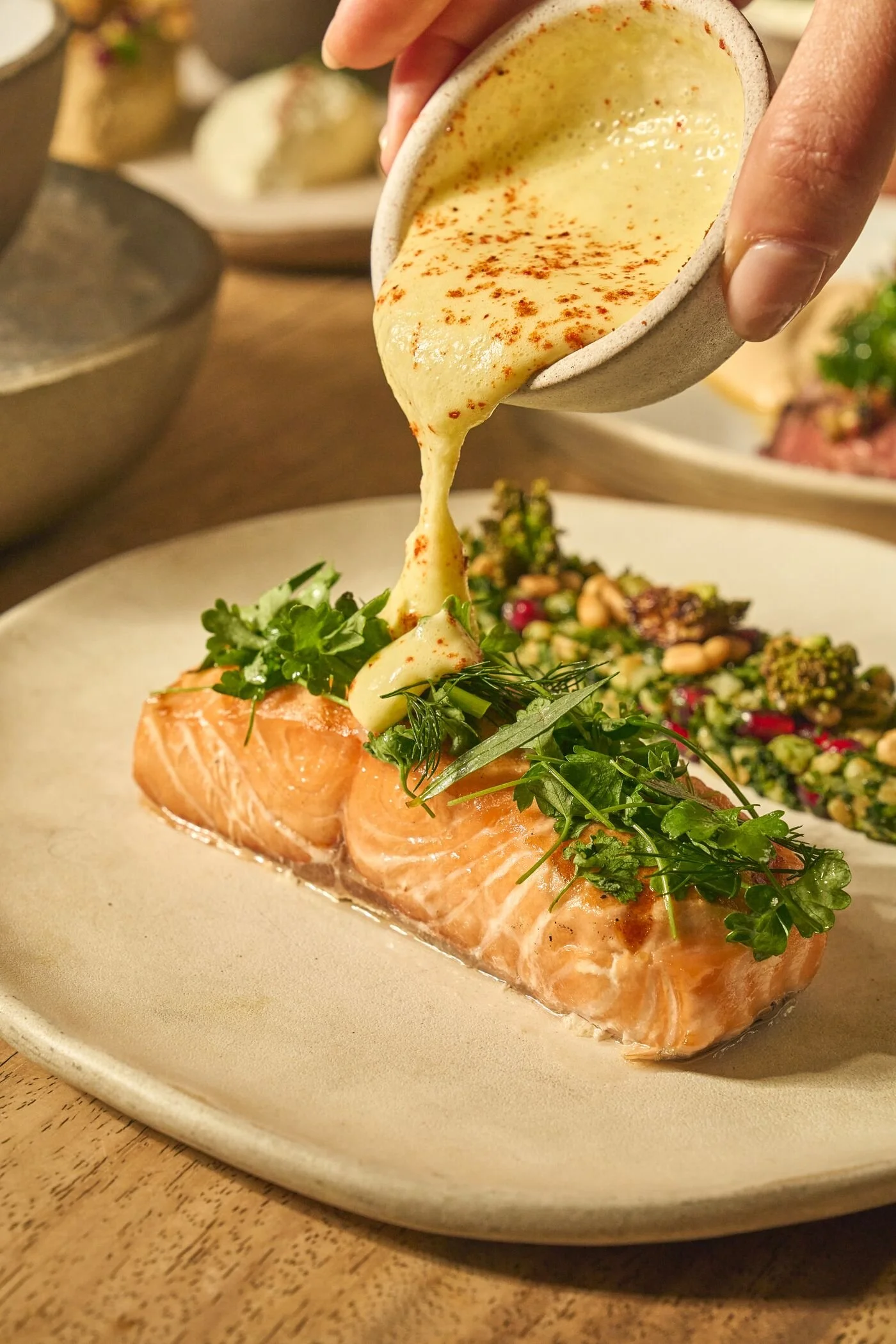 A hand pouring melted cheese over a salmon fillet garnished with green herbs on a beige plate.
