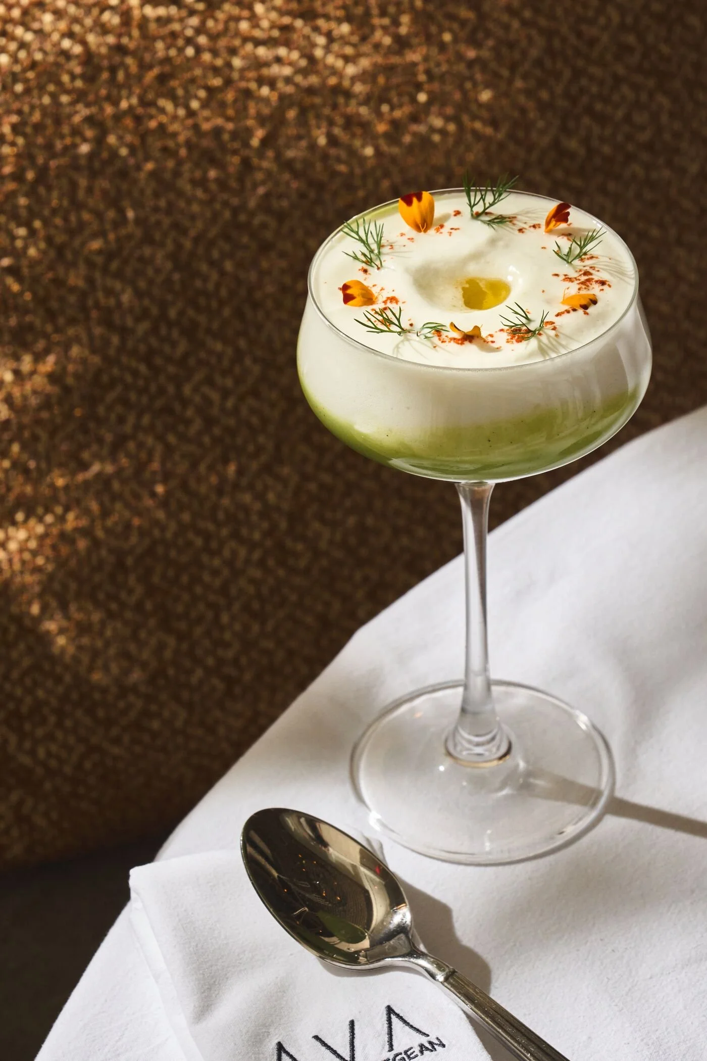 A cocktail glass with a creamy white beverage garnished with orange flower petals, green herbs, and red spice on top, placed on a white tablecloth with a silver spoon nearby.