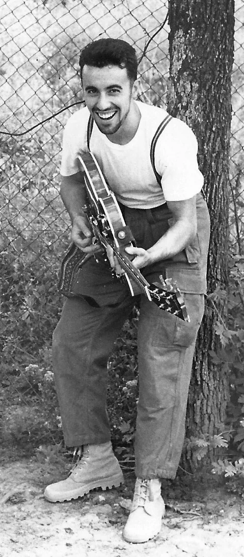 Marco Fazio is playing a guitar outdoors near a tree with a chain-link fence in the background.