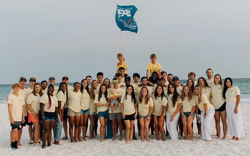 Group of children and teens on a beach, with a person holding a flag and two boys standing on someone's shoulders, all wearing casual beach clothes.