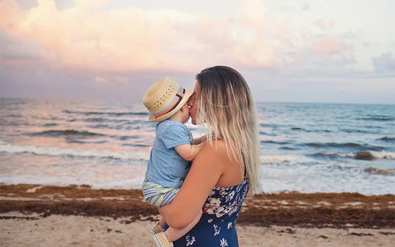 A woman holding a young boy on a beach, both leaning in for a kiss as the sun sets over the ocean.
