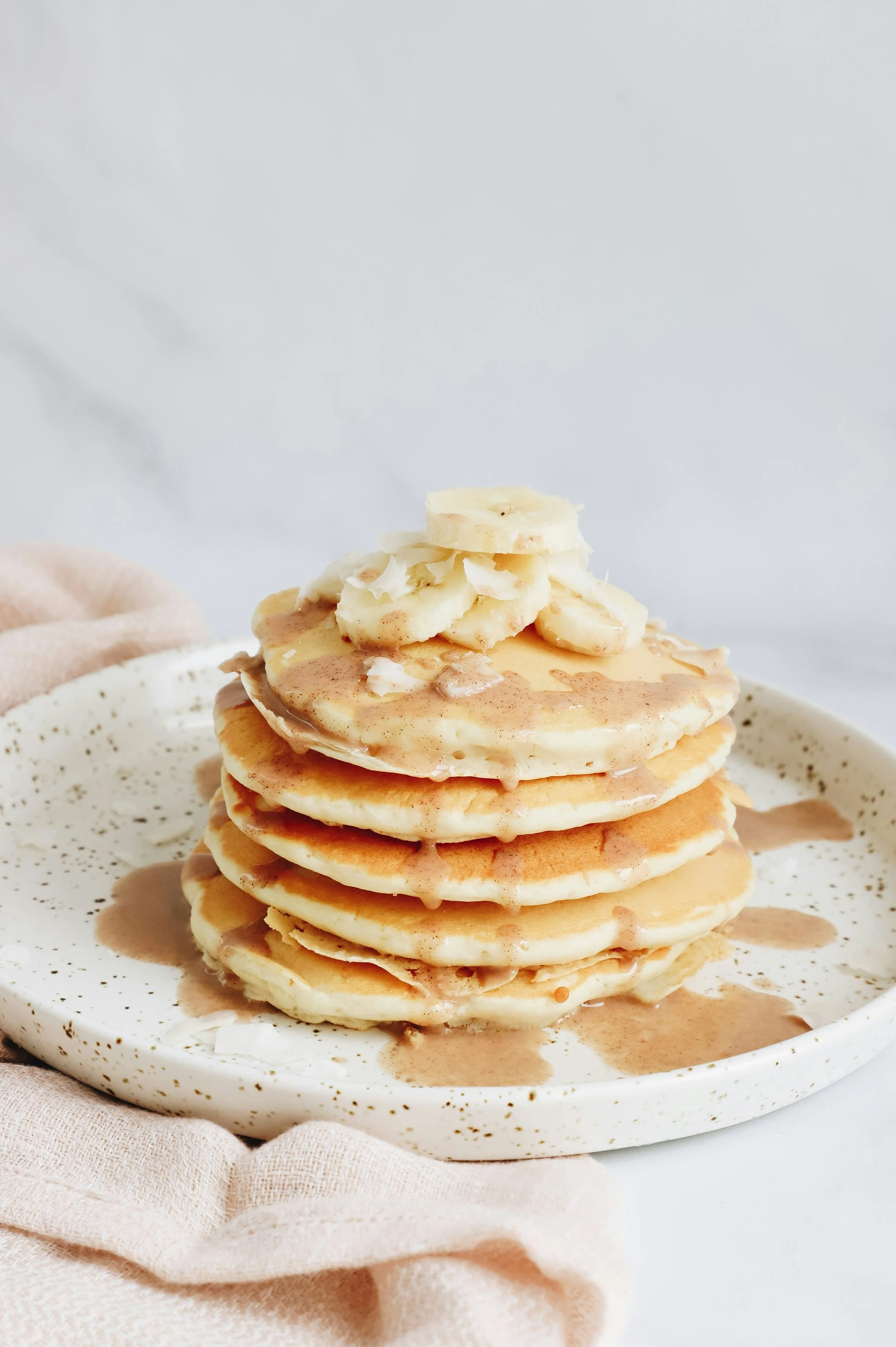 A stack of fluffy pancakes with banana slices and syrup on top, served on a speckled white plate.