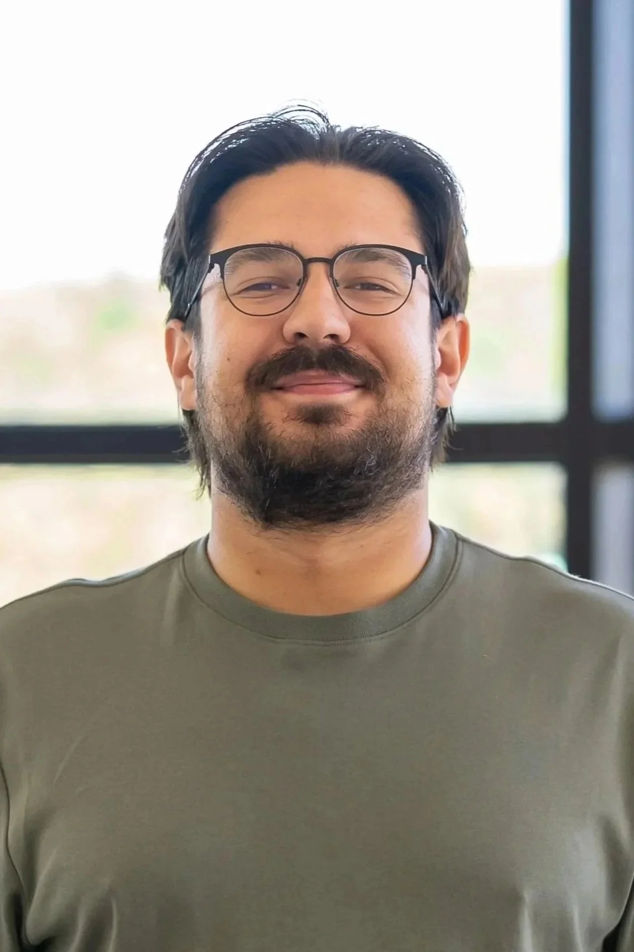 A man with dark hair, glasses, and a beard smiling indoors in front of a window.