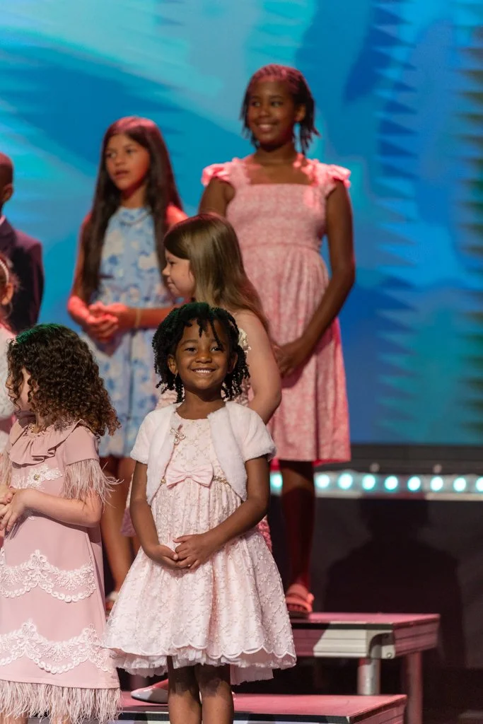 Group of young girls standing on stage, dressed in pink and pastel dresses, smiling during a performance or event.